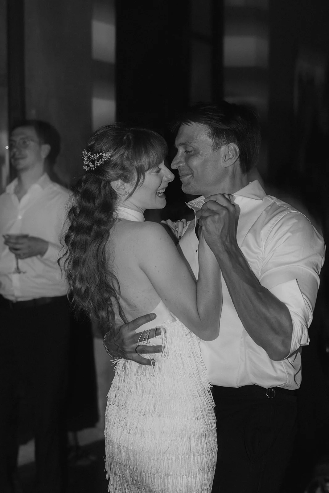 Ein Paar tanzt eng auf einer Hochzeit, sie lächeln sich verliebt an, er trägt ein weißes Hemd, sie trägt ein weißes Kleid mit Fransen.