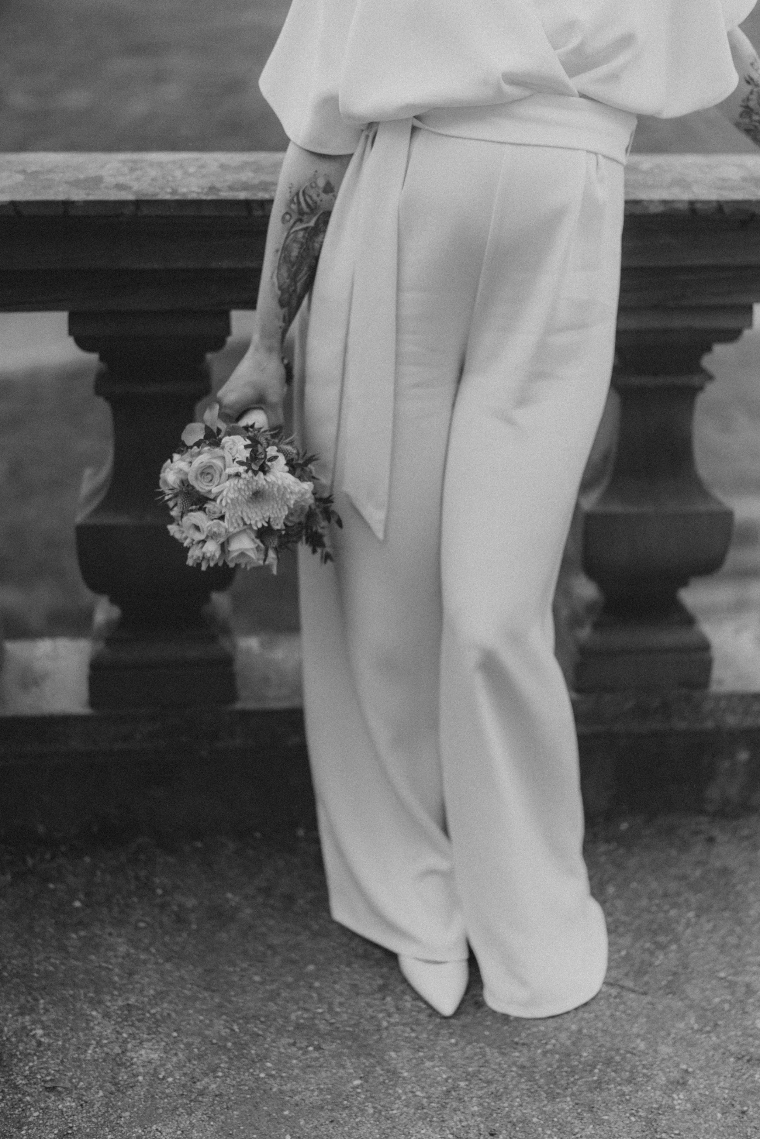 Schwarz-Weiß-Foto einer Person in weißen, weiten Hosen und einer Bluse, die einen Blumenstrauß hält, vor einem Stein-Tisch.