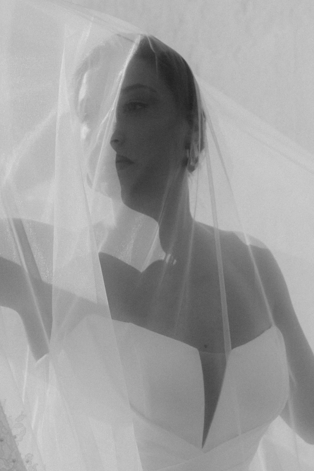 Schwarz-Weiß-Foto einer jungen Frau mit glattem Haar, die hinter einem transparenten Stoff steht, der ein Kleid oder eine Decke zu sein scheint.