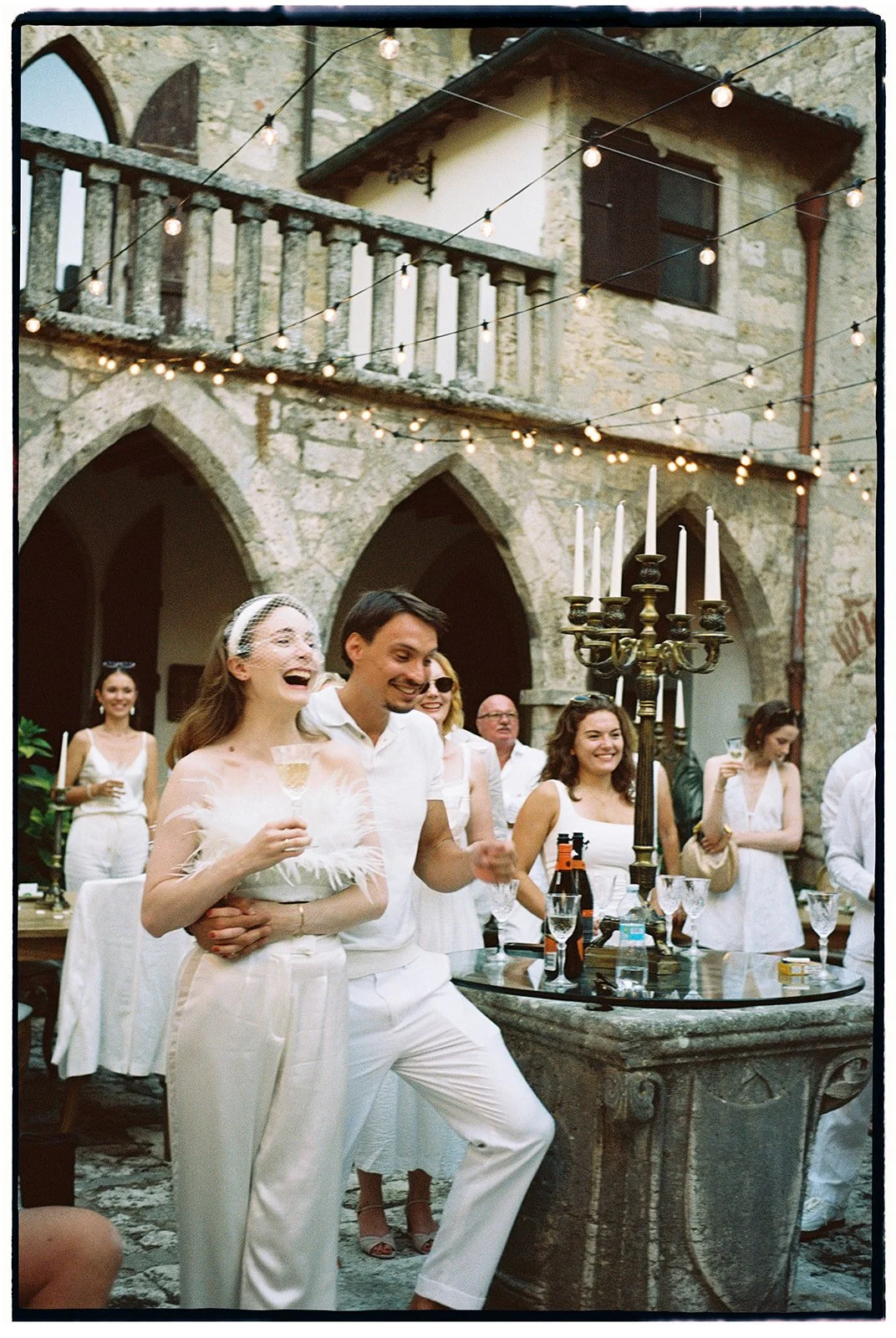 Menschen feiern eine Party im Innenhof eines alten Steingebäudes mit Lichterketten und Kerzen.