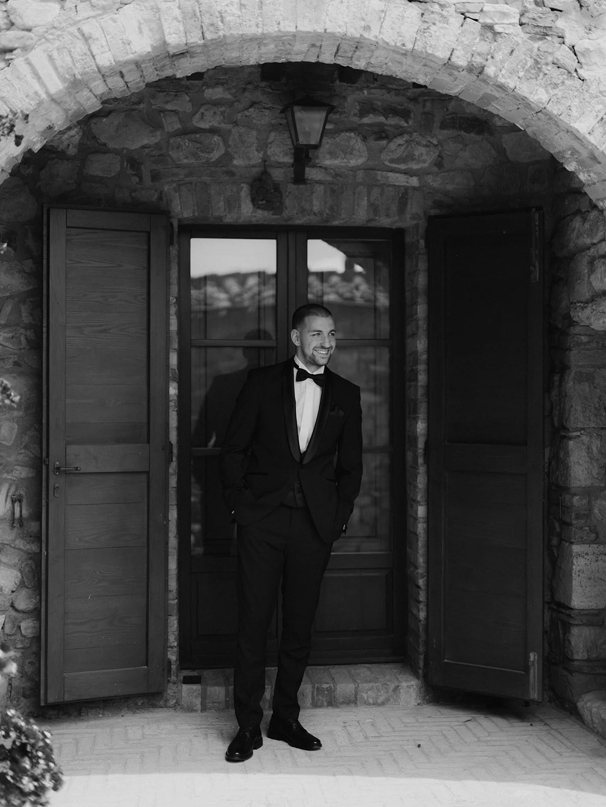 Bräutigam  in formeller Abendgarderobe, schwarze Anzug, Fliege, steht vor einer Holztür in einer Steingebäude, lächelt, Schwarz-Weiß-Foto, auf Poggio Covili in der Toskana bei einer destinationwedding