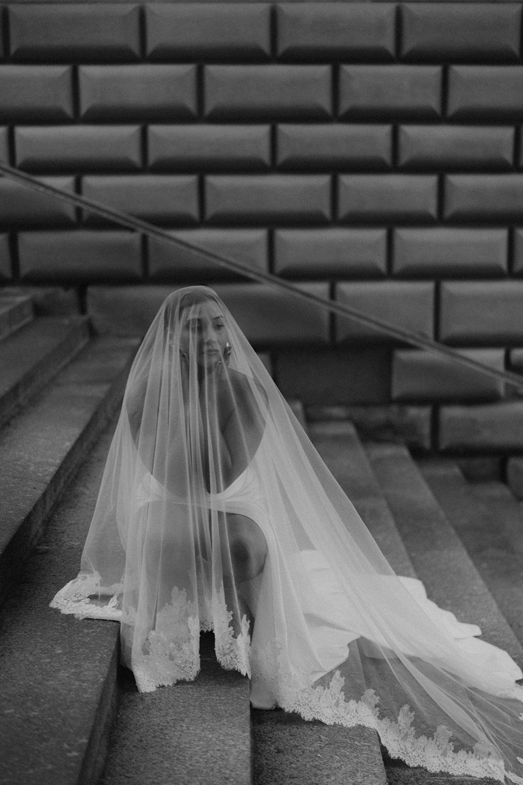 Schwarz-weiß-Fotografie einer jungen Frau mit einem Brautkleid, die auf Treppen sitzt, bedeckt von einem durchsichtigen Schleier.