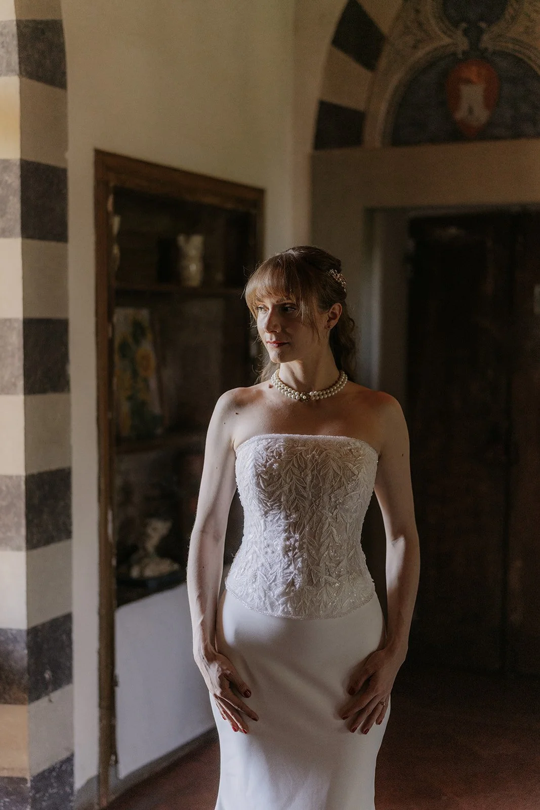 Eine Frau trägt ein elegantes weißes Brautkleid mit Stickerei und Perlenkette in einem Raum mit dunklen Wänden und einer gemalten Wappen im Hintergrund.