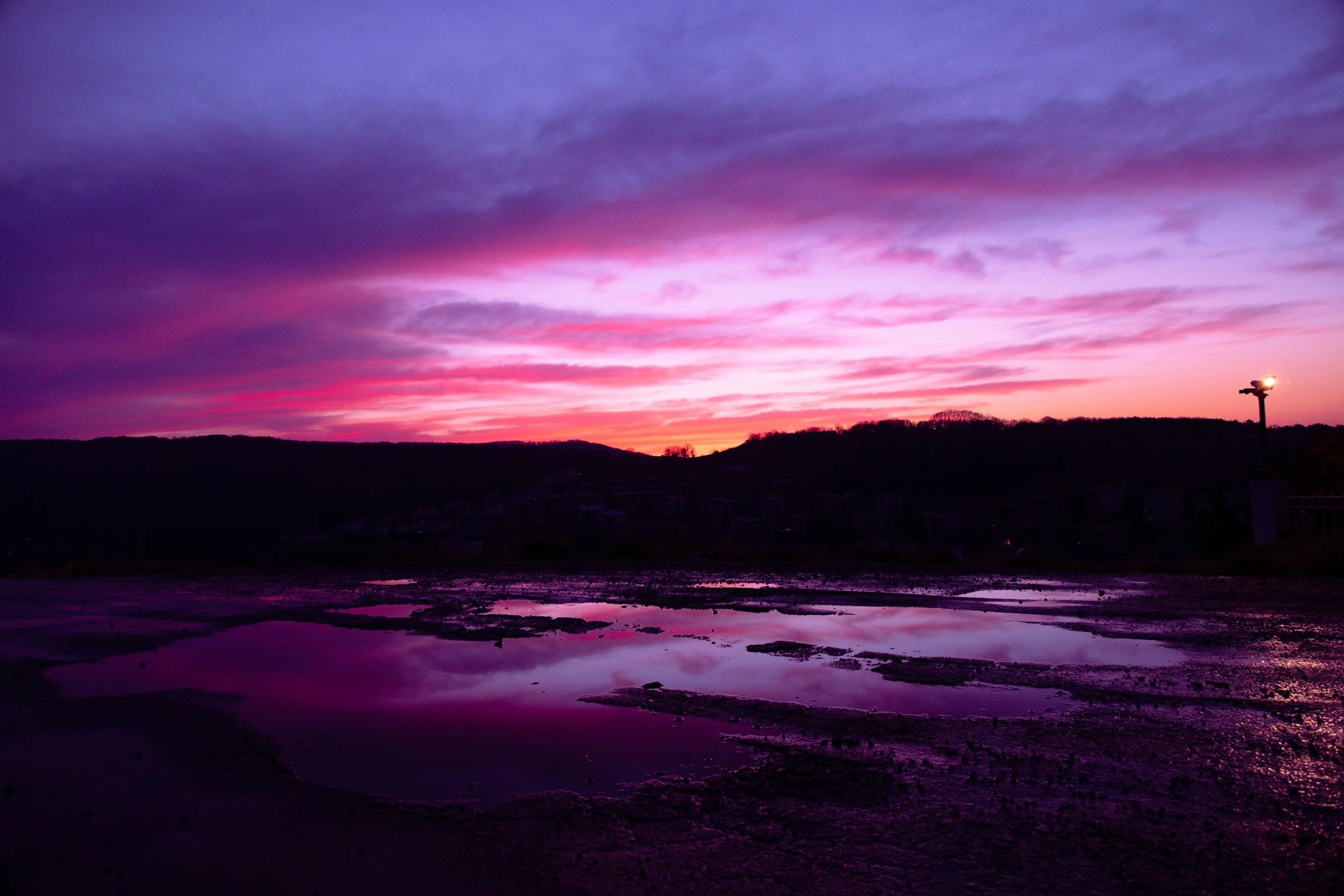 Colorful sunset over a landscape with puddles of water reflecting the sky, hills in the distance, and a single streetlamp on the right.