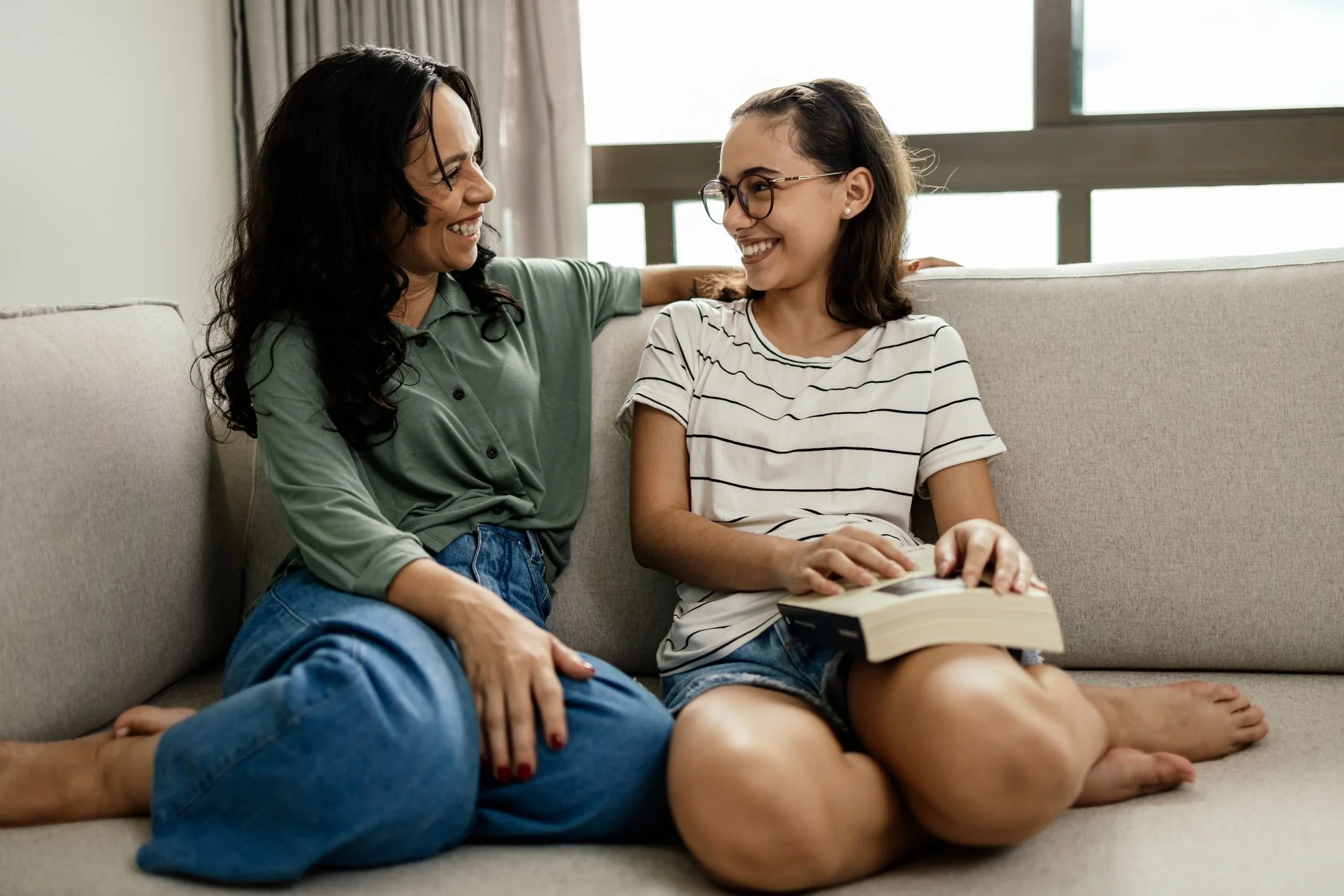 A woman and a young girl sitting on a beige couch, smiling at each other, with the girl holding a book.