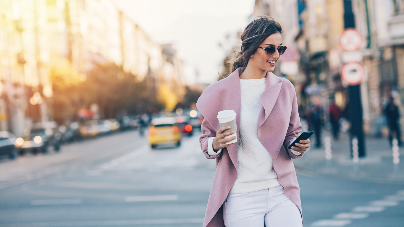 A woman in a pink coat and sunglasses walking in an urban street, holding a coffee cup in one hand and a smartphone in the other.