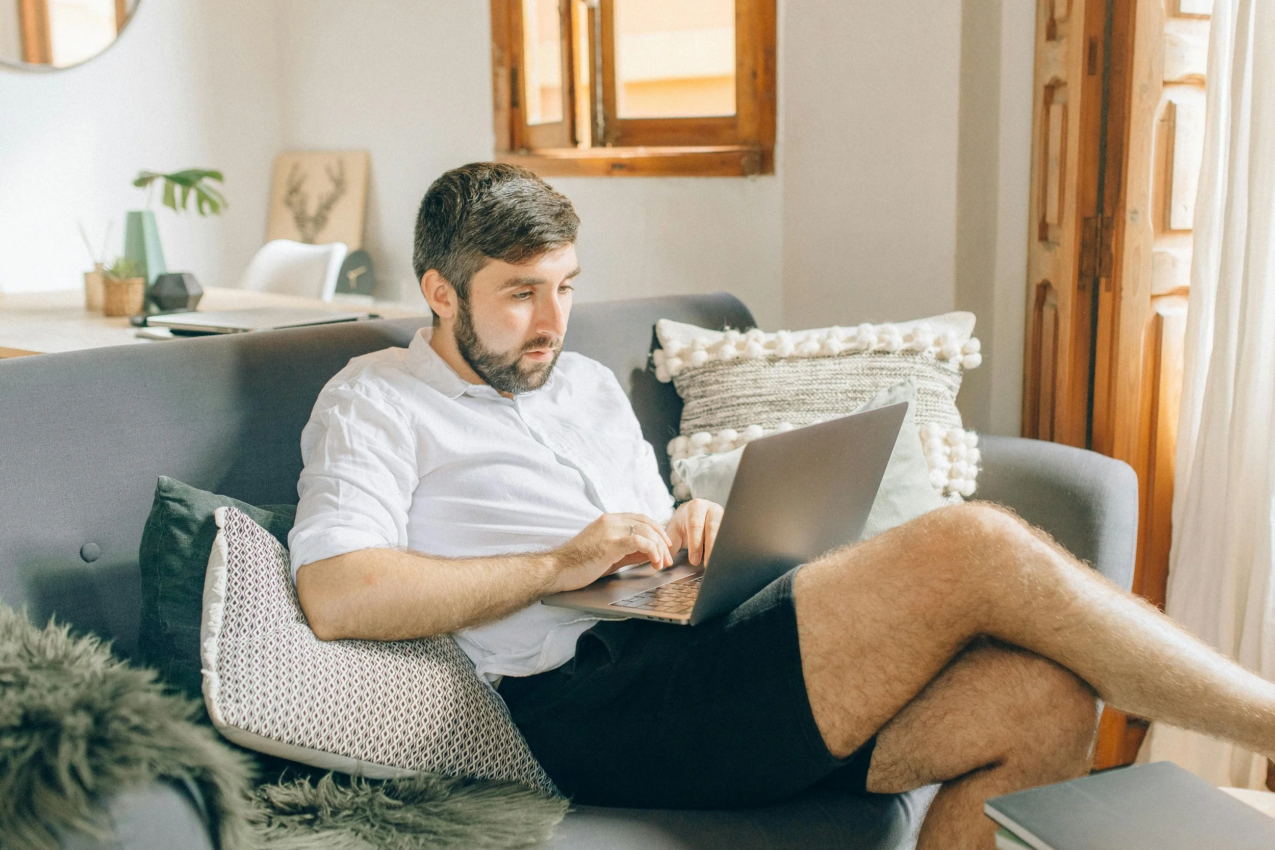 A man with a beard wearing a white shirt and dark shorts sitting on a sofa with a laptop.