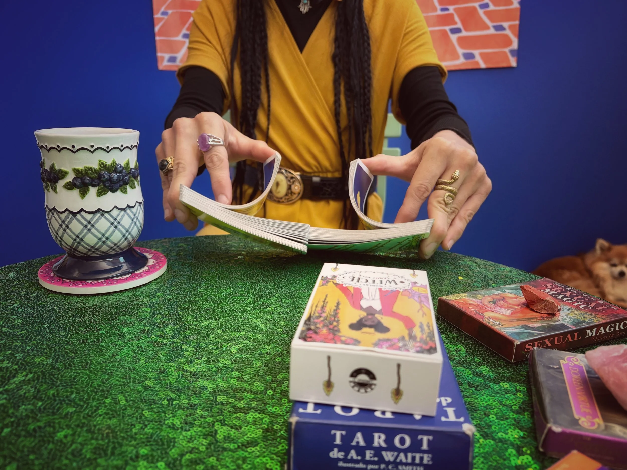 A person wearing a yellow dress with a black long sleeve shirt underneath is flipping through a deck of tarot cards on a green sequined tablecloth. There are various tarot card boxes, including one labeled 'Tarot de A.E. Waite' and another labeled 'Sexual Magic.' A decorative mug with a blueberry and leaf pattern sits on a pink coaster to the left. In the background, there is a blue wall with a brick pattern. A small dog is partially visible lying on the right side of the image.