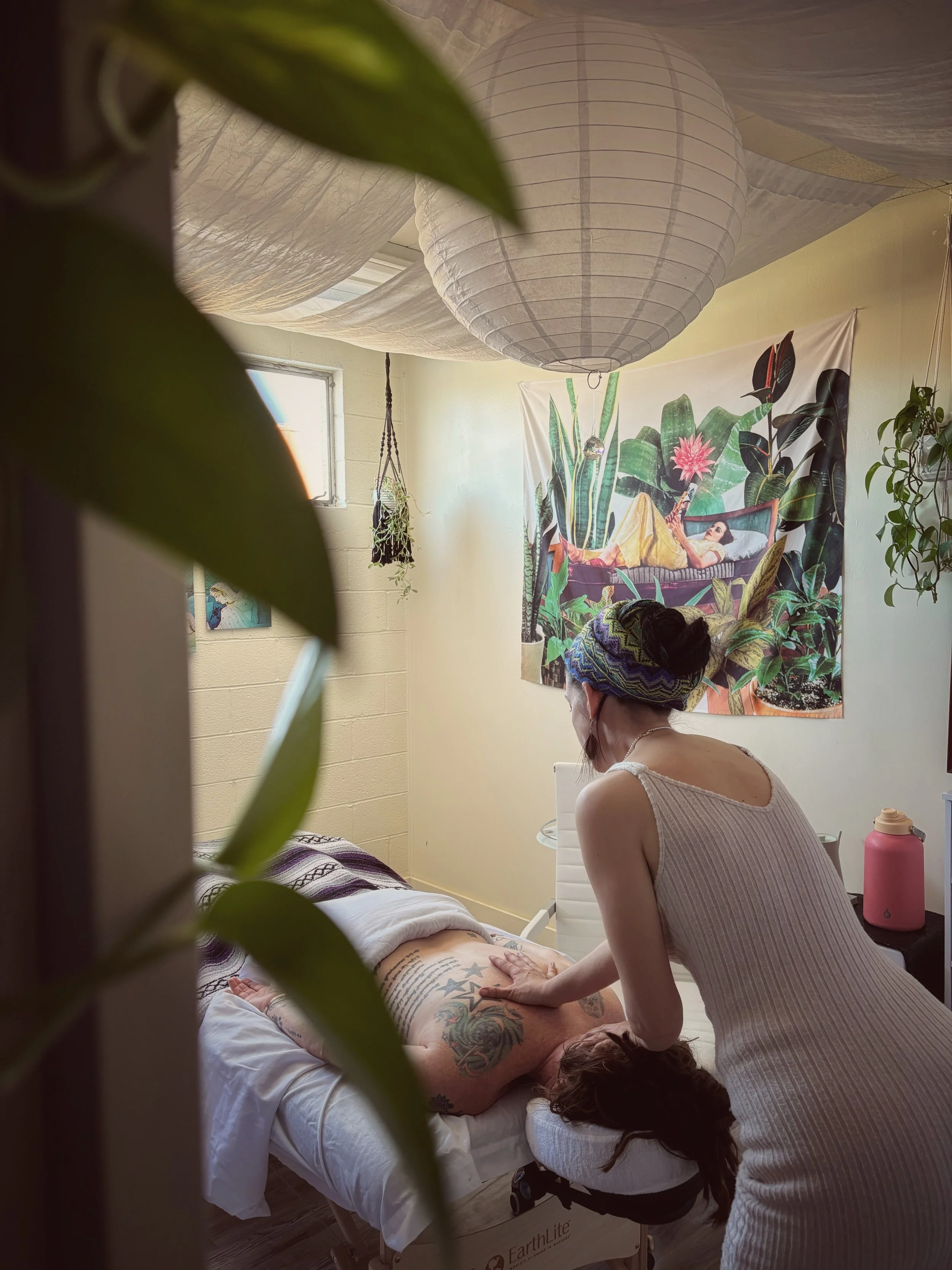 A woman receiving a massage from a massage therapist in a room decorated with plants and a large tapestry of a woman lounging among tropical plants.