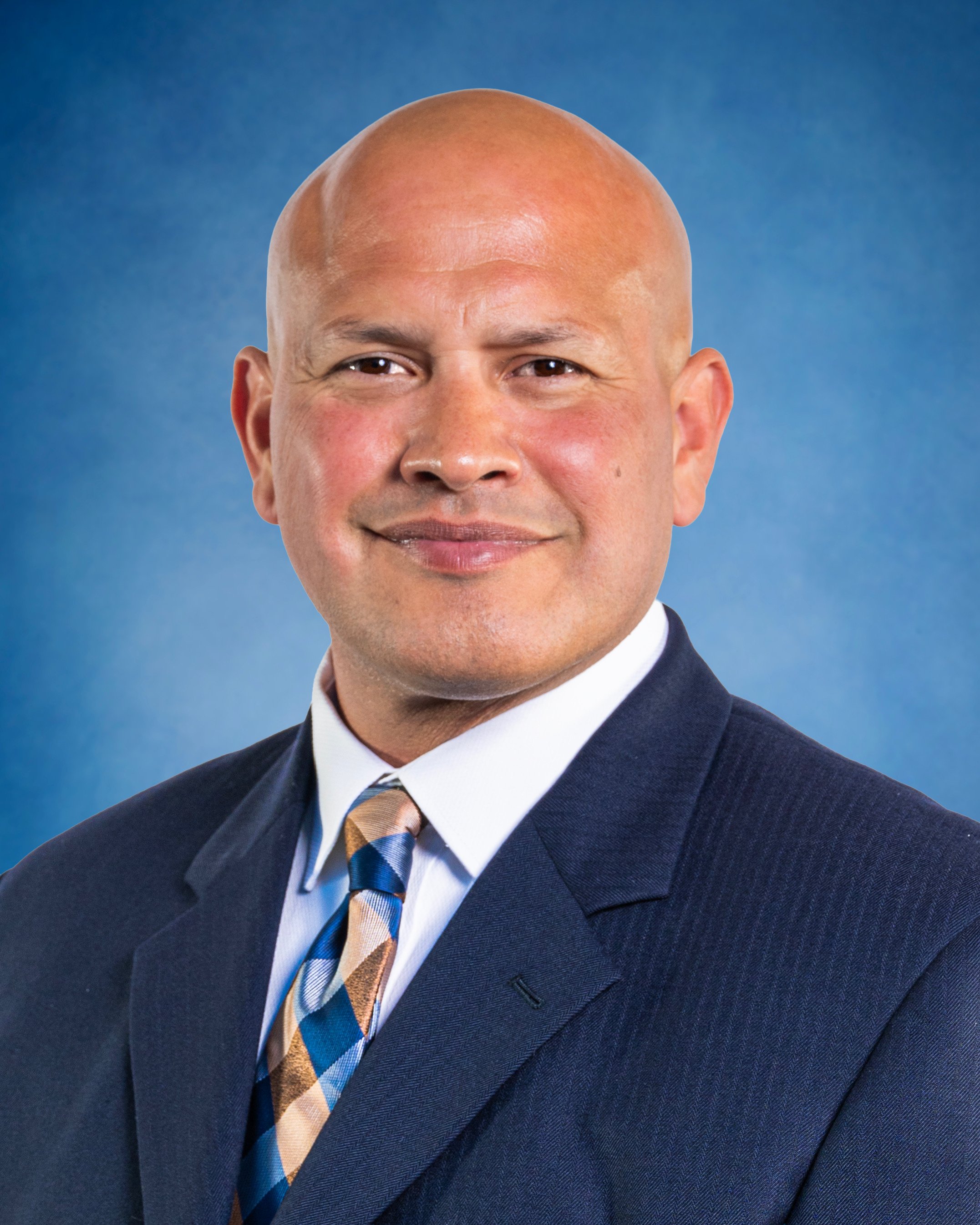 Professional headshot of Luis Lara in a dark business suit with a white shirt and a patterned tie, smiling against a blue background.