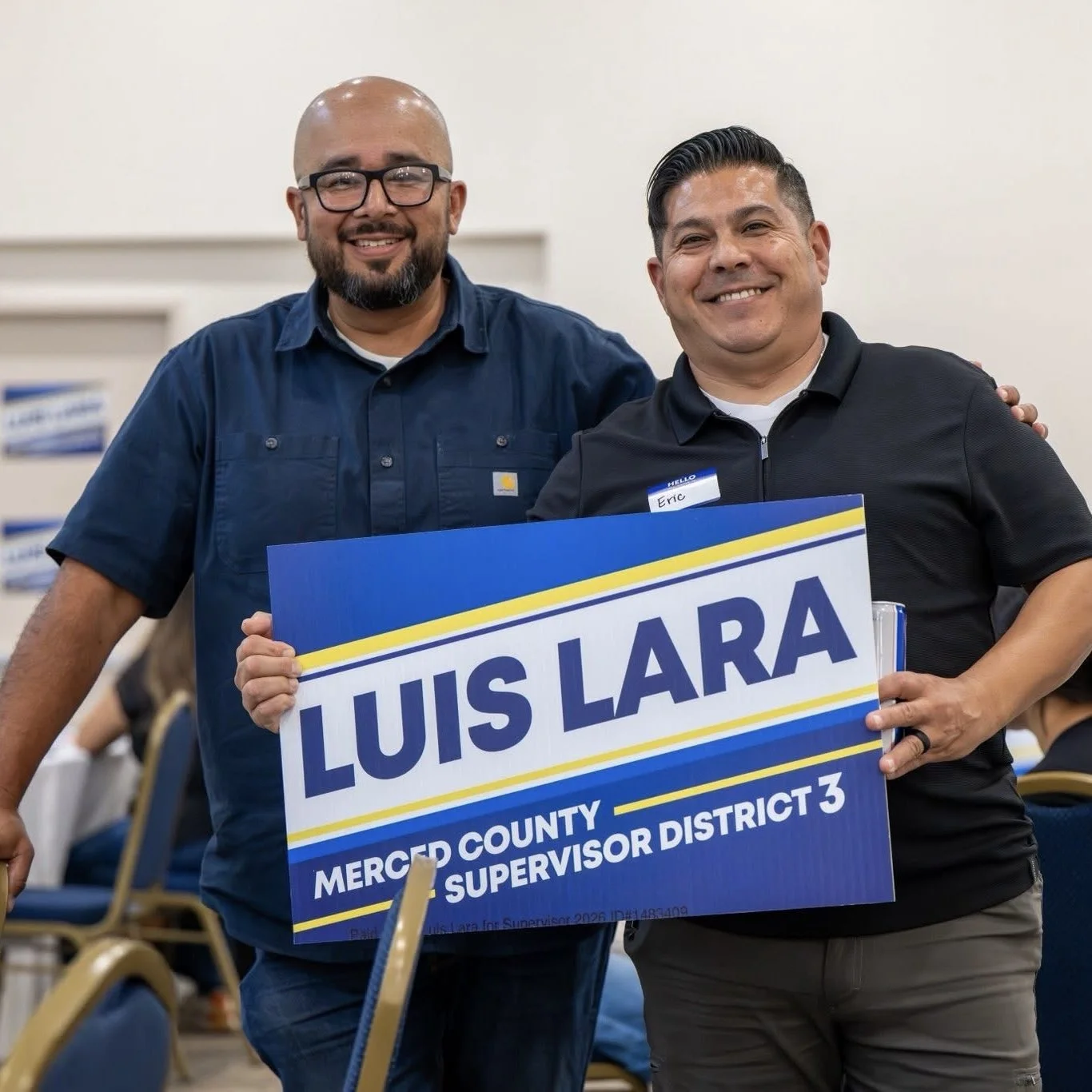 Two men smiling and holding a sign for Luis Lara, a county supervisor candidate in Merced County District 3, during a political event.