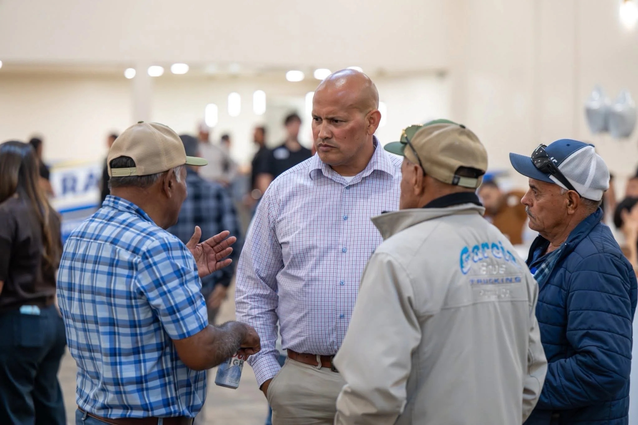 Luis Lara engaged in conversation with three men at an indoor event, with one man wearing a purple checkered shirt and the others in casual jackets and hats, with a crowd and some balloons visible in the background.