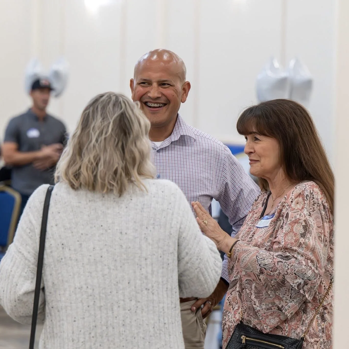 Luis Lara and two women engaged in conversation, smiling, in a well-lit indoor setting, with a blurred person in the background.