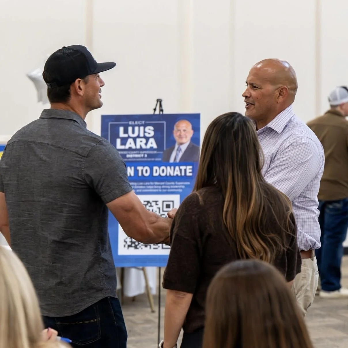 Luis Lara and a man shaking hands at a political event, with a woman standing nearby, in front of a Luis Lara campaign sign.