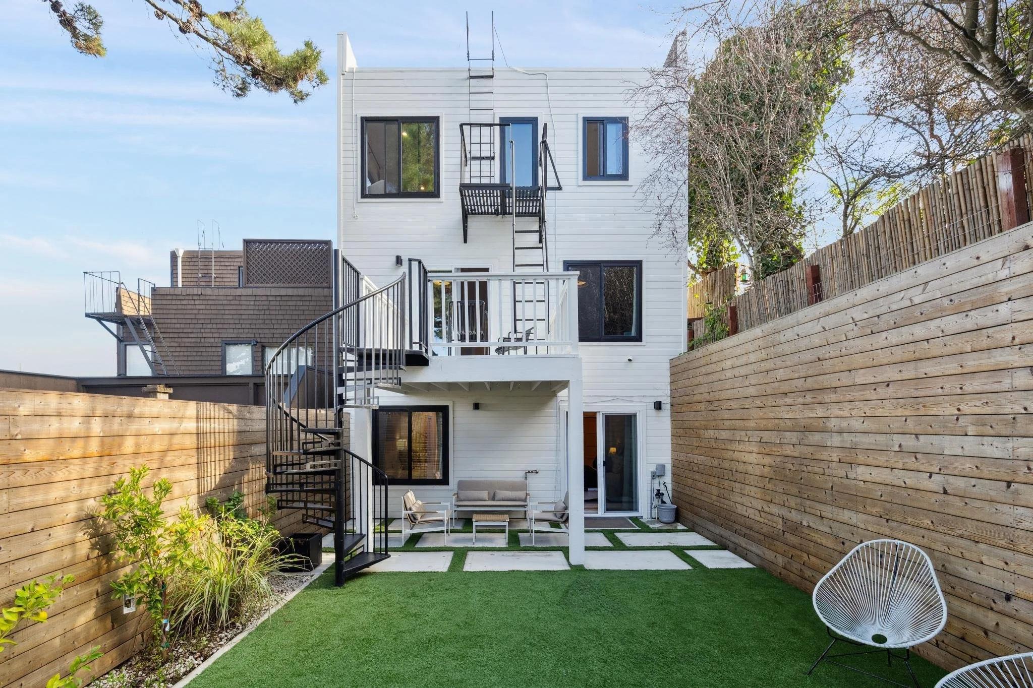 View of a modern, multi-story house with white siding, black-framed windows, and an outdoor patio area enclosed by wooden fencing. The yard features green artificial turf, patio furniture, and a spiral staircase leading to the upper balcony.
