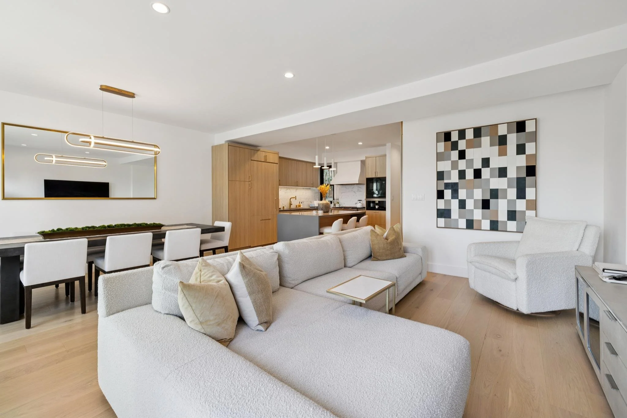 Bright, modern living room with white sectional sofa, white armchair, and wooden flooring. The room features a large wall mirror, abstract geometric wall art, and an open-concept kitchen in the background with wooden cabinets, a white hood, and a kit