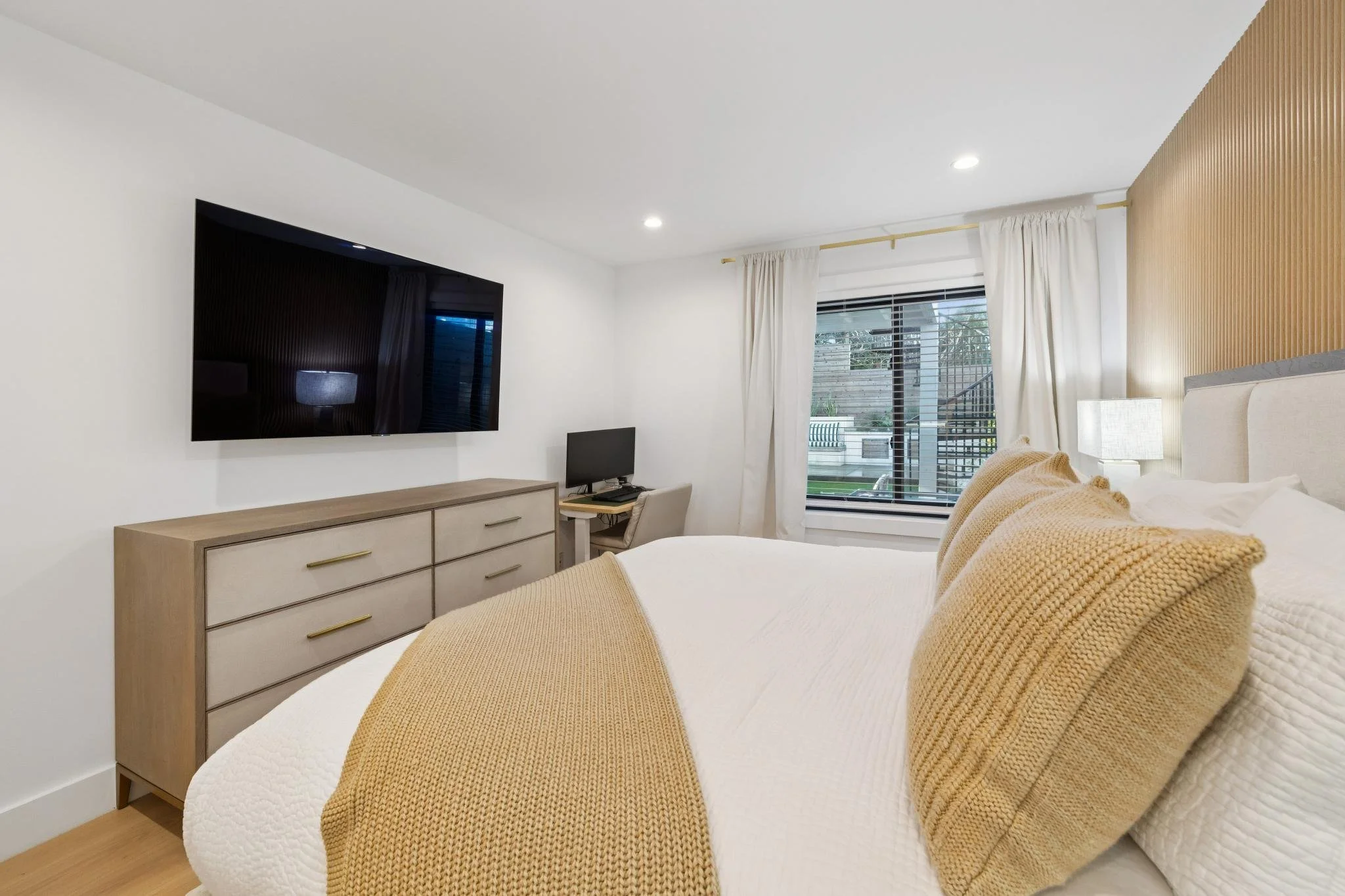 A modern bedroom with a bed, beige pillows, a bedside lamp, a dresser, a television, a window with blinds, and a small workspace.