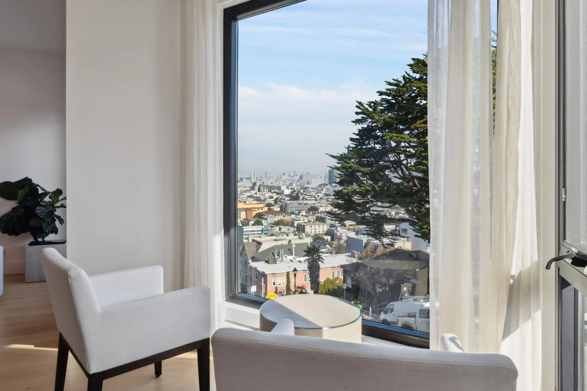 A view from inside a modern apartment with white chairs and a side table, looking out through large glass windows at a cityscape with many buildings and a large tree outside.