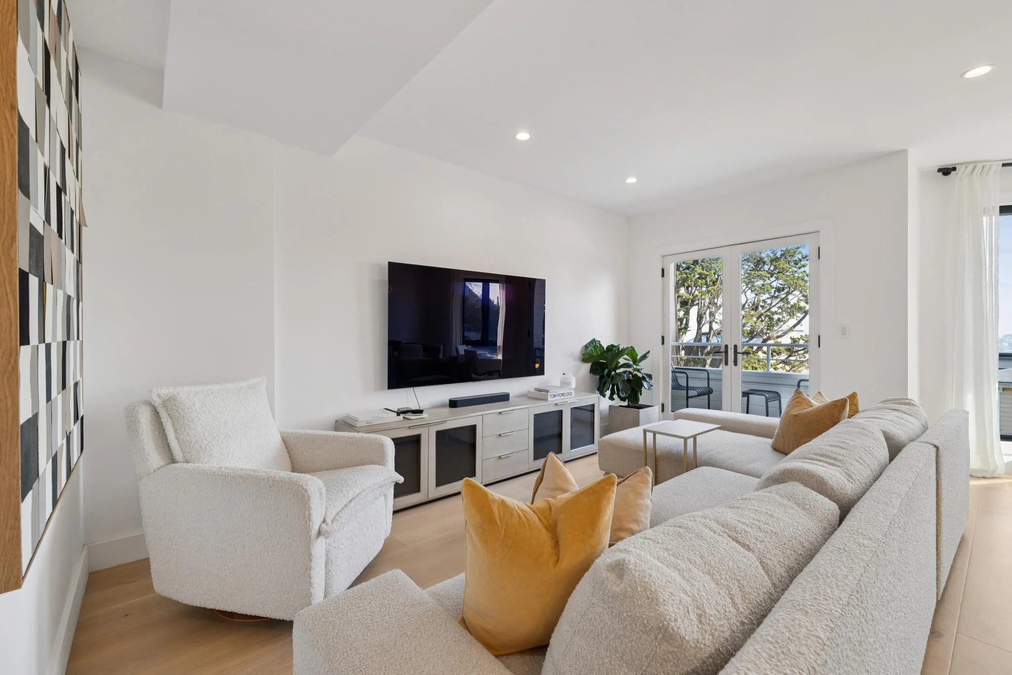 Living room with white walls and furniture, large flat-screen TV on a white entertainment center, cream sofa with gold pillows, white armchair, glass doors leading to a balcony with outdoor chairs, and a large green plant.