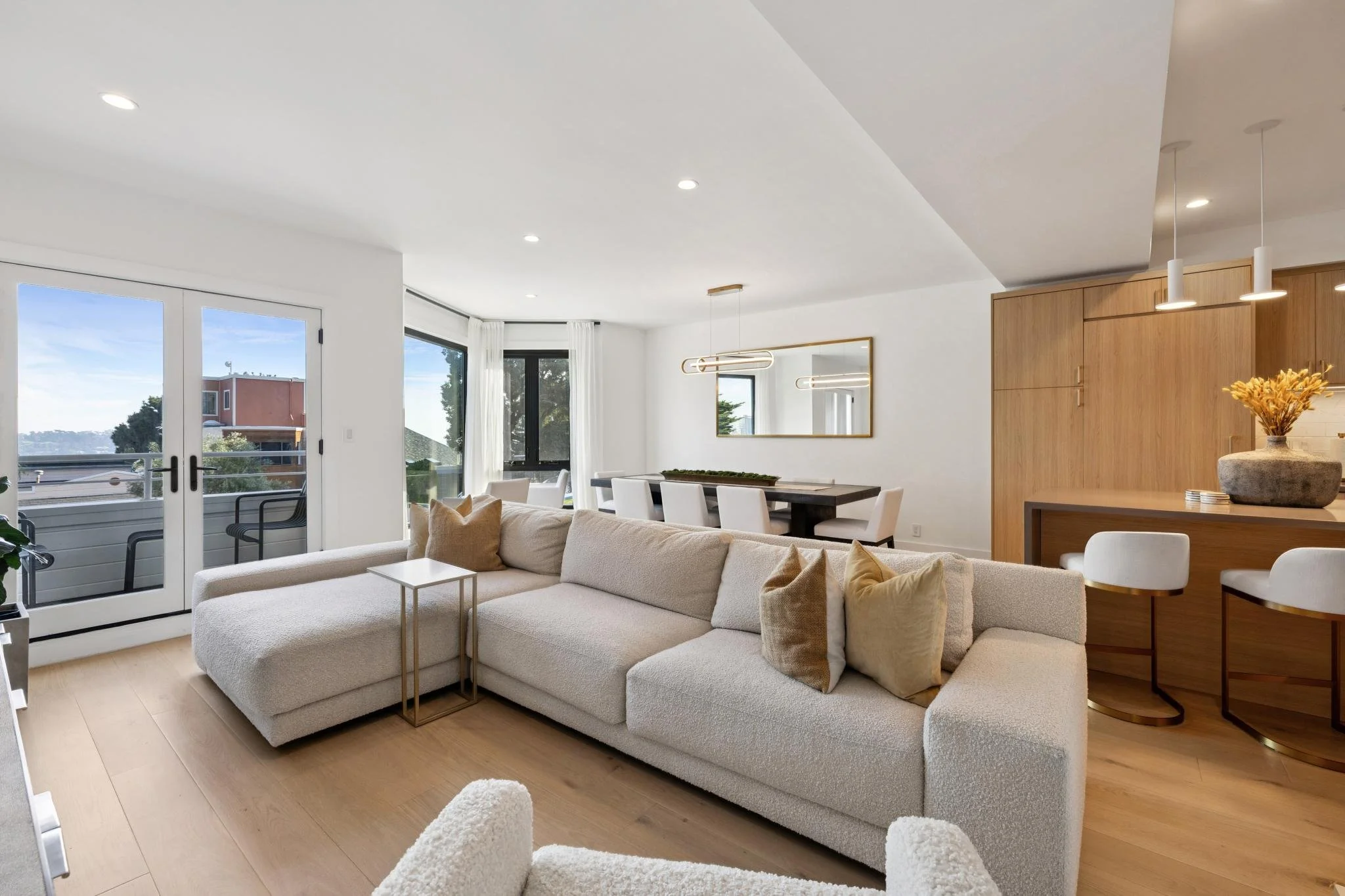 Bright, modern living room with white sofa, beige pillows, light wooden flooring, and large glass doors leading to an outdoor balcony. Dining area with white chairs and a black table, and a wooden kitchen area with white bar stools.