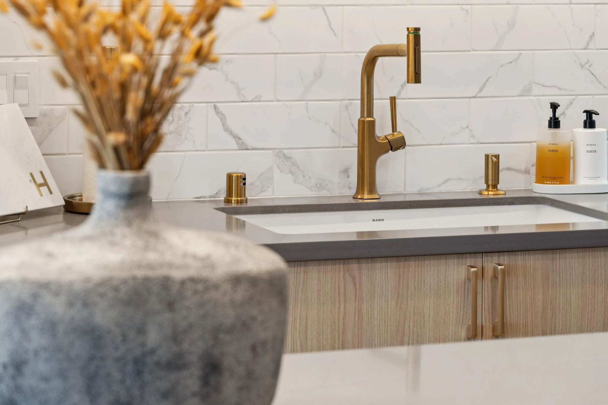 Modern kitchen sink with gold faucet and soap dispensers, marble backsplash, and wooden cabinets.