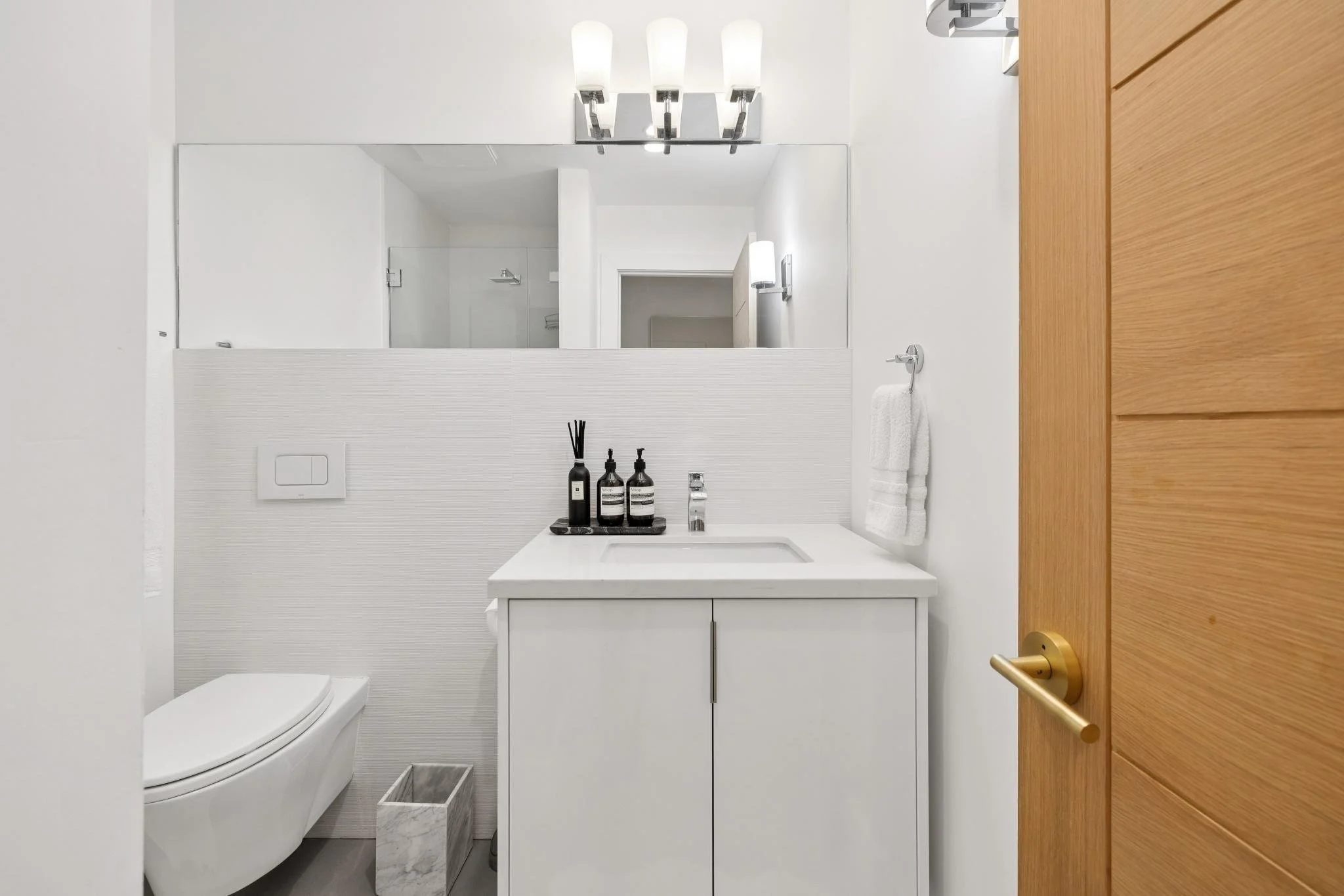 Modern bathroom with white walls, a sink with a white cabinet, a large mirror, and a wooden door. Bathroom accessories include black bottles with reed diffuser, soap, and lotion on the sink, a towel on a wall hook, and a toilet with a marble trash bi