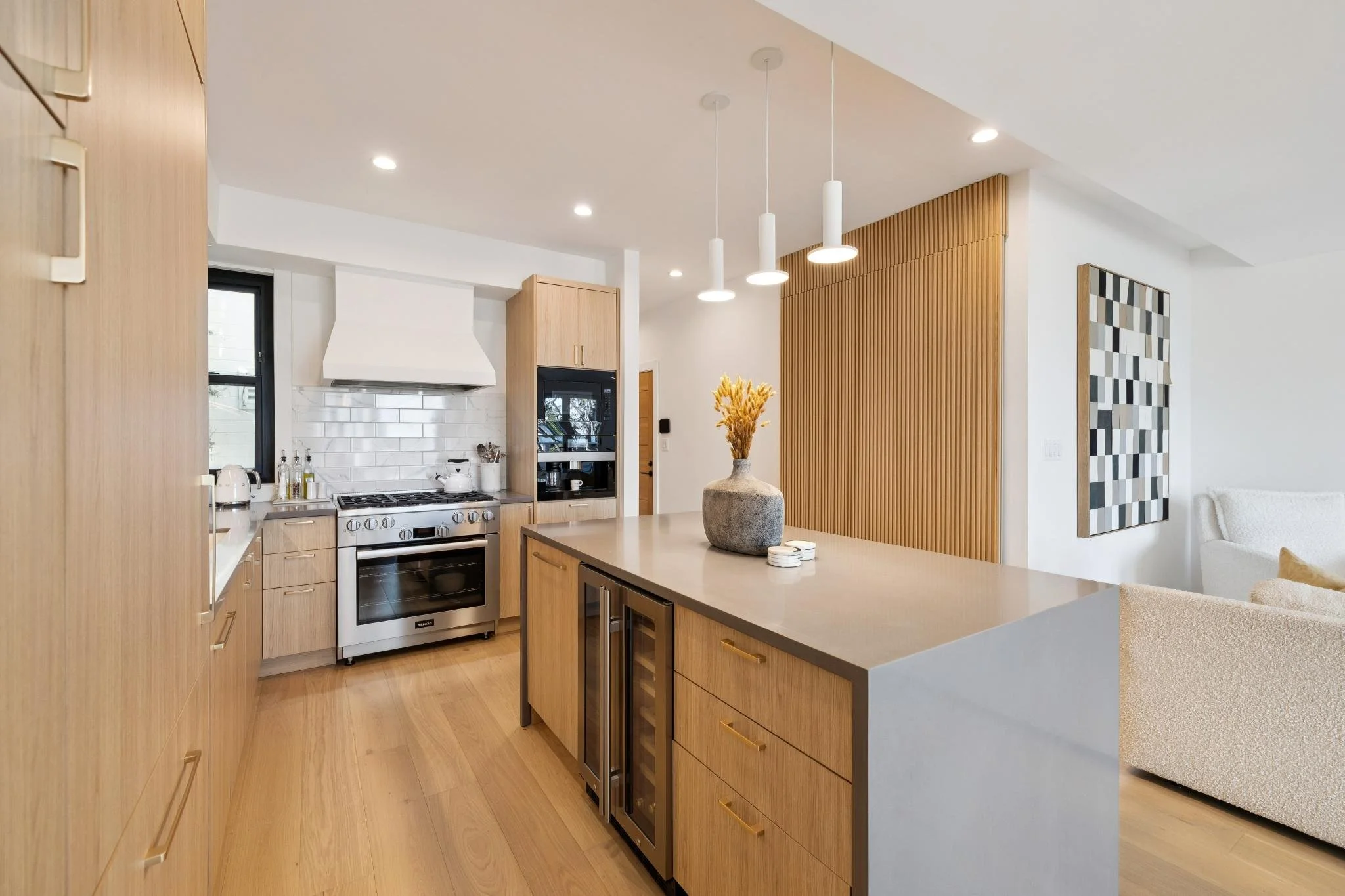 Modern kitchen with light wood cabinets, a center island with a light gray countertop, a stainless steel oven, white subway tile backsplash, and decorative pendant lights. Decor includes a vase with dried flowers on the island.
