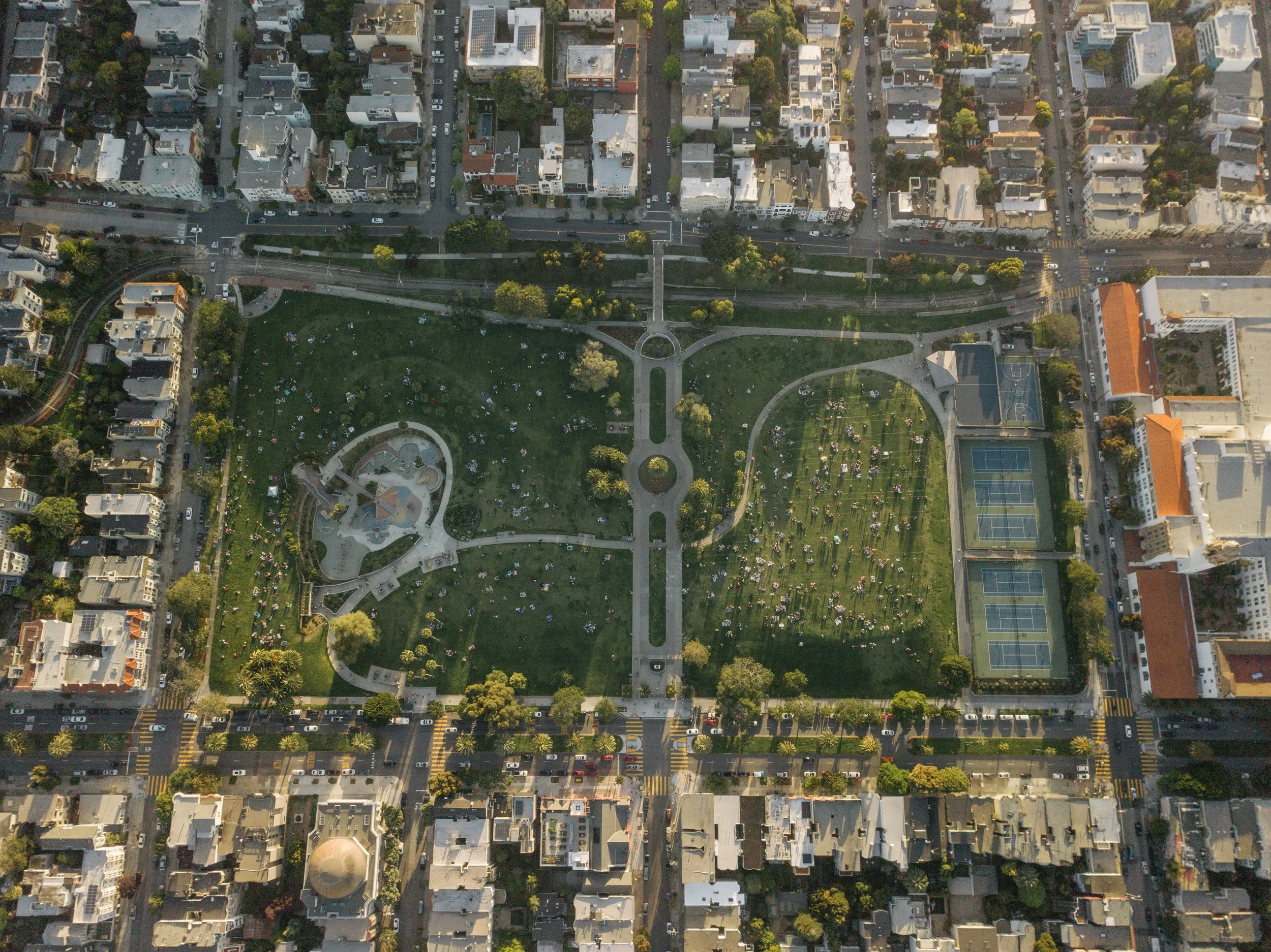 An aerial view of a city park surrounded by streets and buildings, with a circular fountain, pathways, tennis courts, and many park visitors.