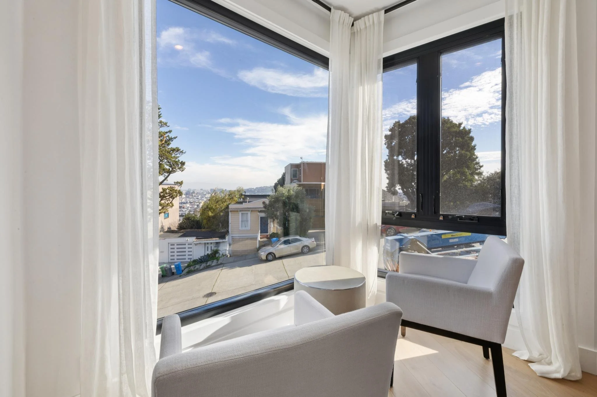 A bright corner with two white armchairs, a small round side table, large windows with sheer white curtains, and a view of a neighborhood street with parked cars and houses under a partly cloudy sky.