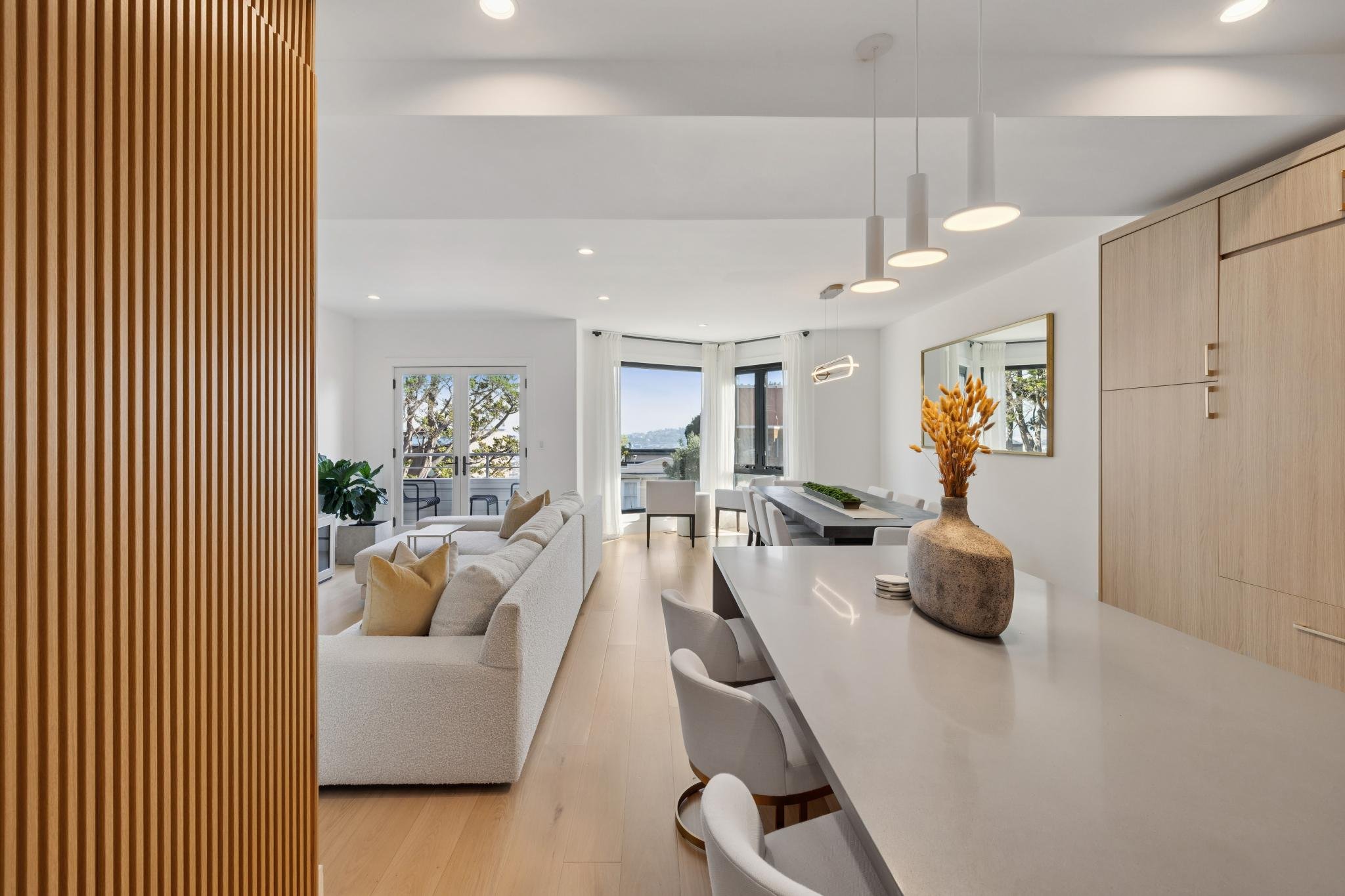 Modern living room with white sofa, dining table with chairs, and large windows with curtains, illuminated with ceiling lights and natural daylight.