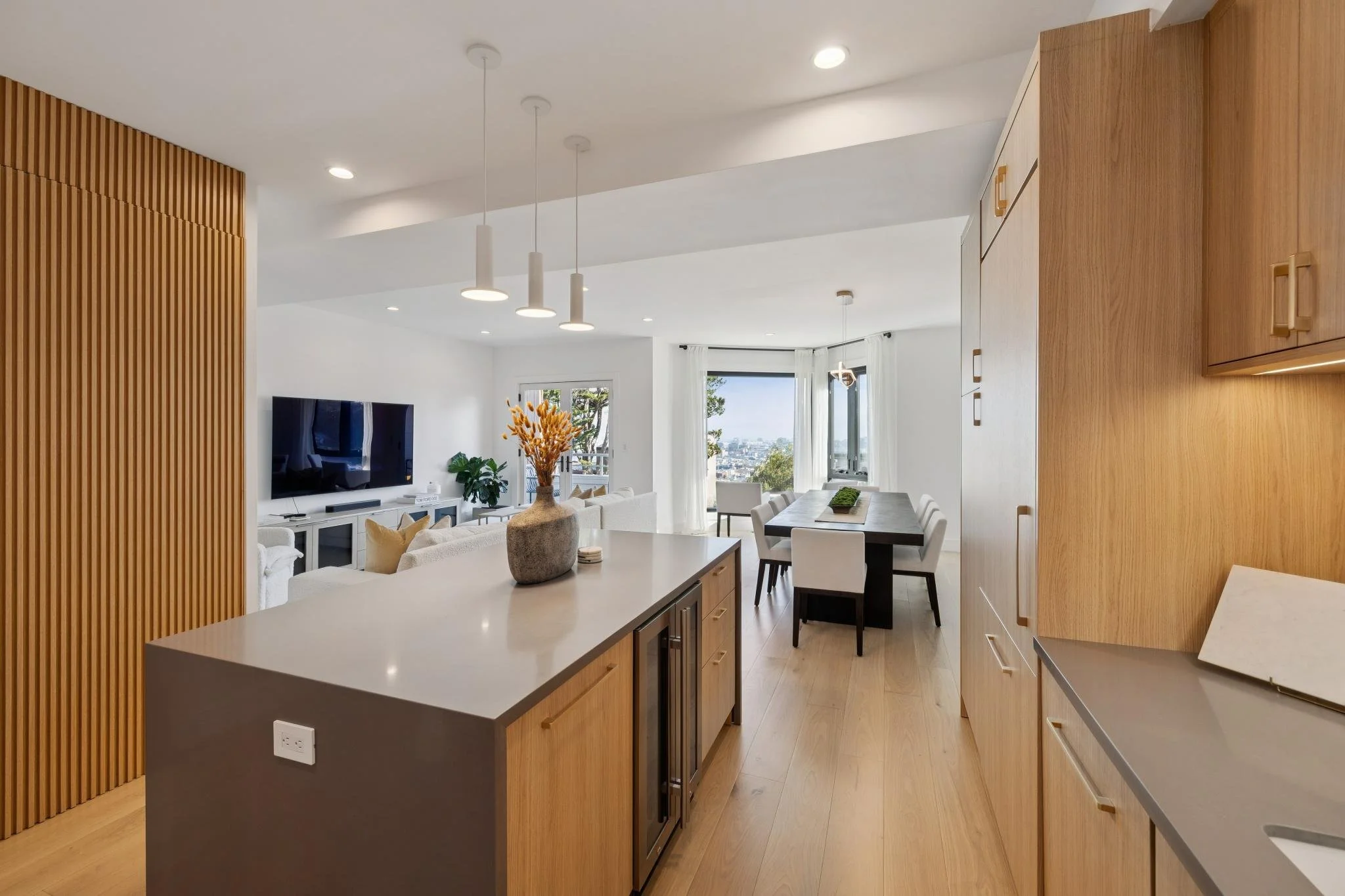Modern open-concept living room and kitchen with wooden accents, white furniture, and a view of the city through large windows.