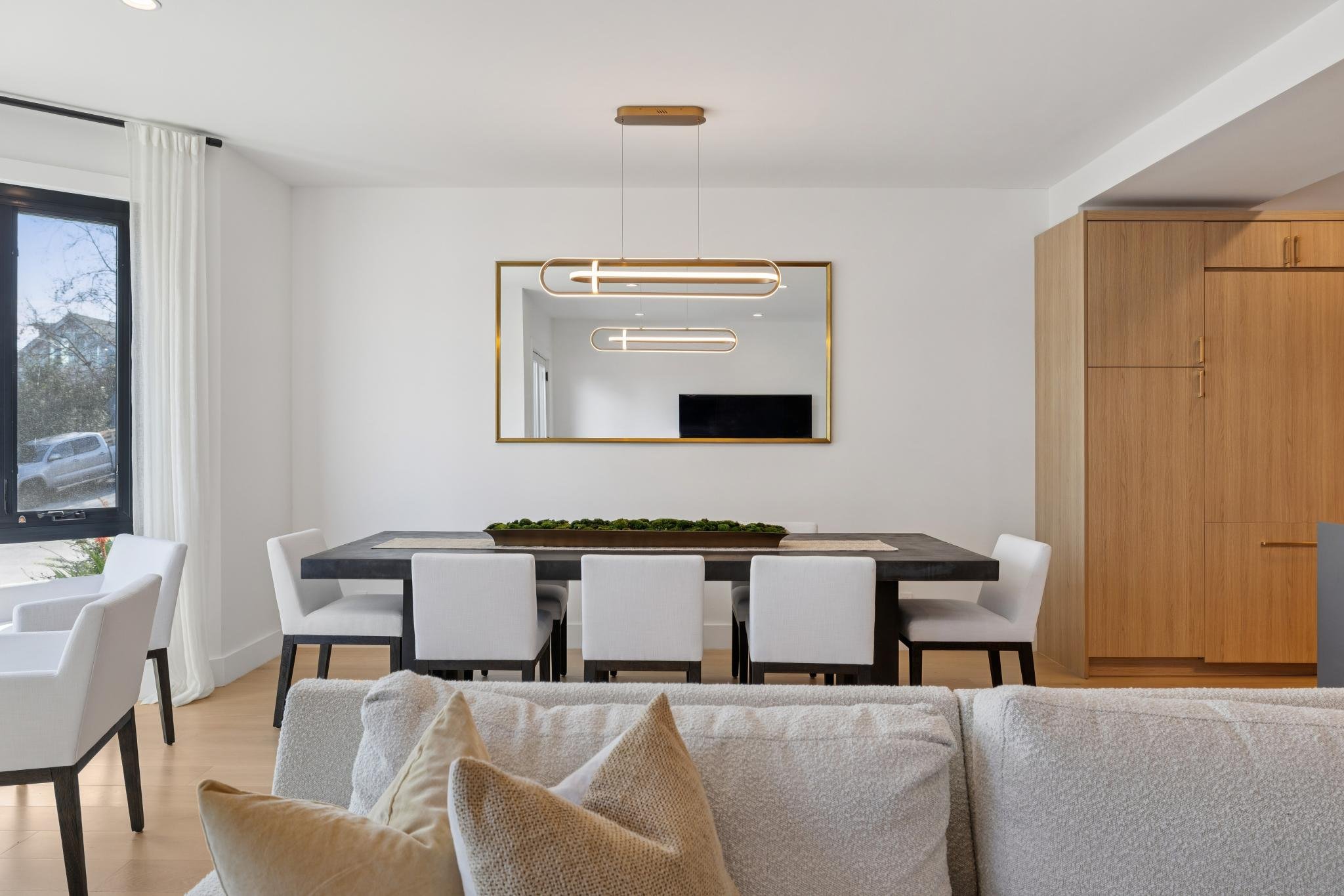 Modern dining room with a black table, white chairs, a large mirror with a gold frame, and a wooden cabinet.