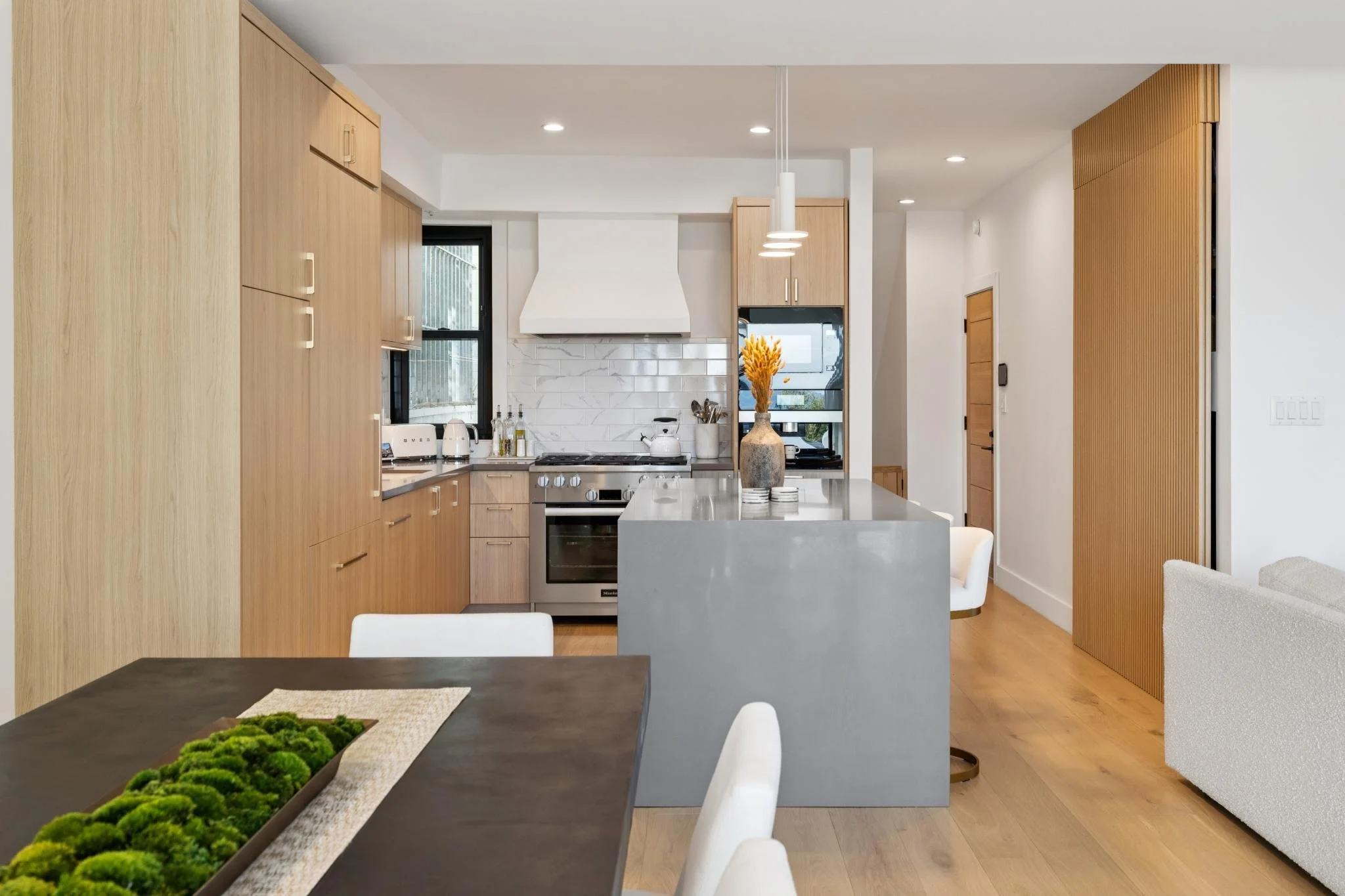 Modern kitchen with wooden cabinets, gray island, white backsplash, and black-framed windows, with dining table in foreground.