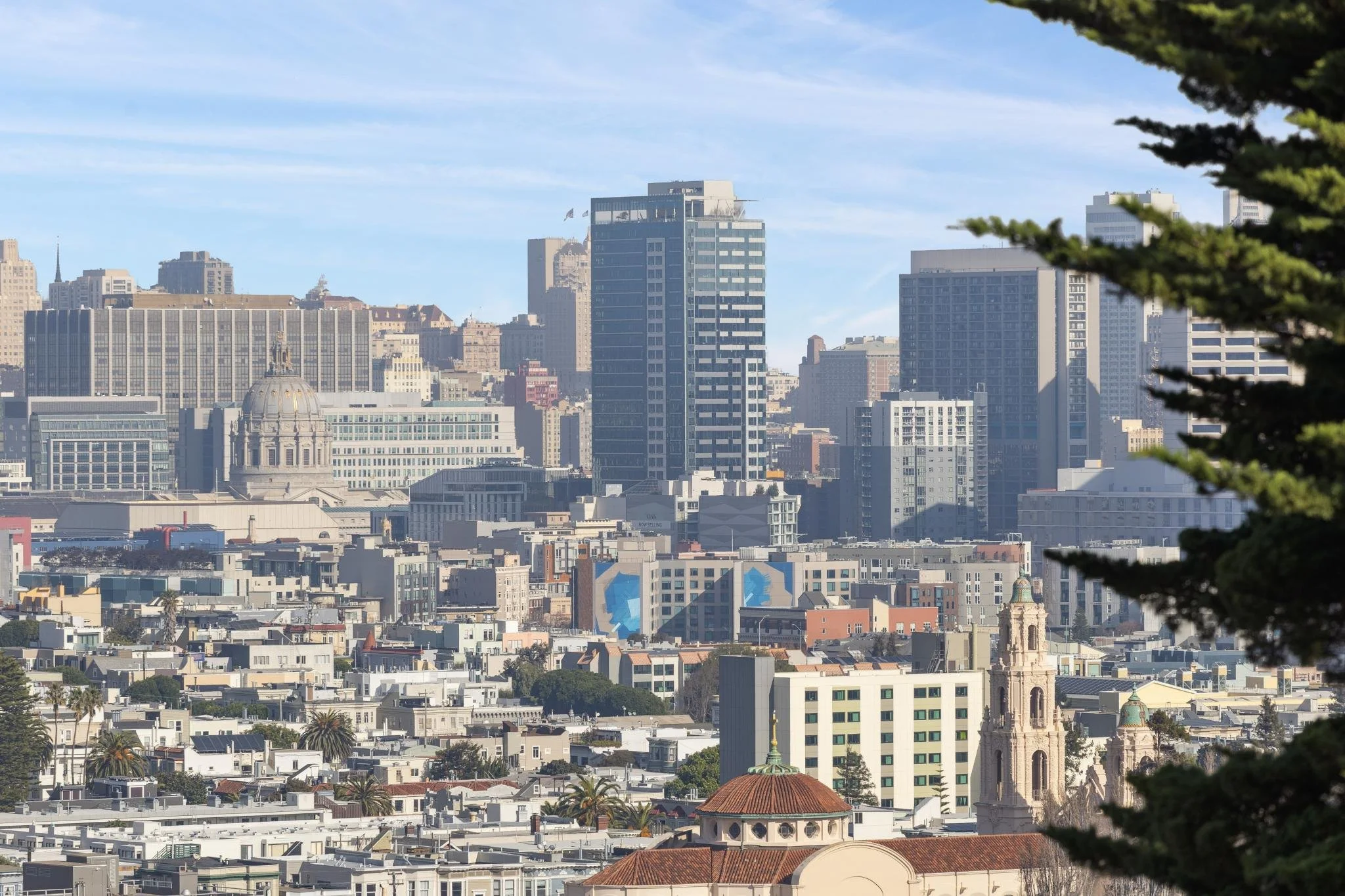 City skyline with various tall buildings and skyscrapers, some with distinctive architectural features, under a partly cloudy sky, with tree branches on the right side.