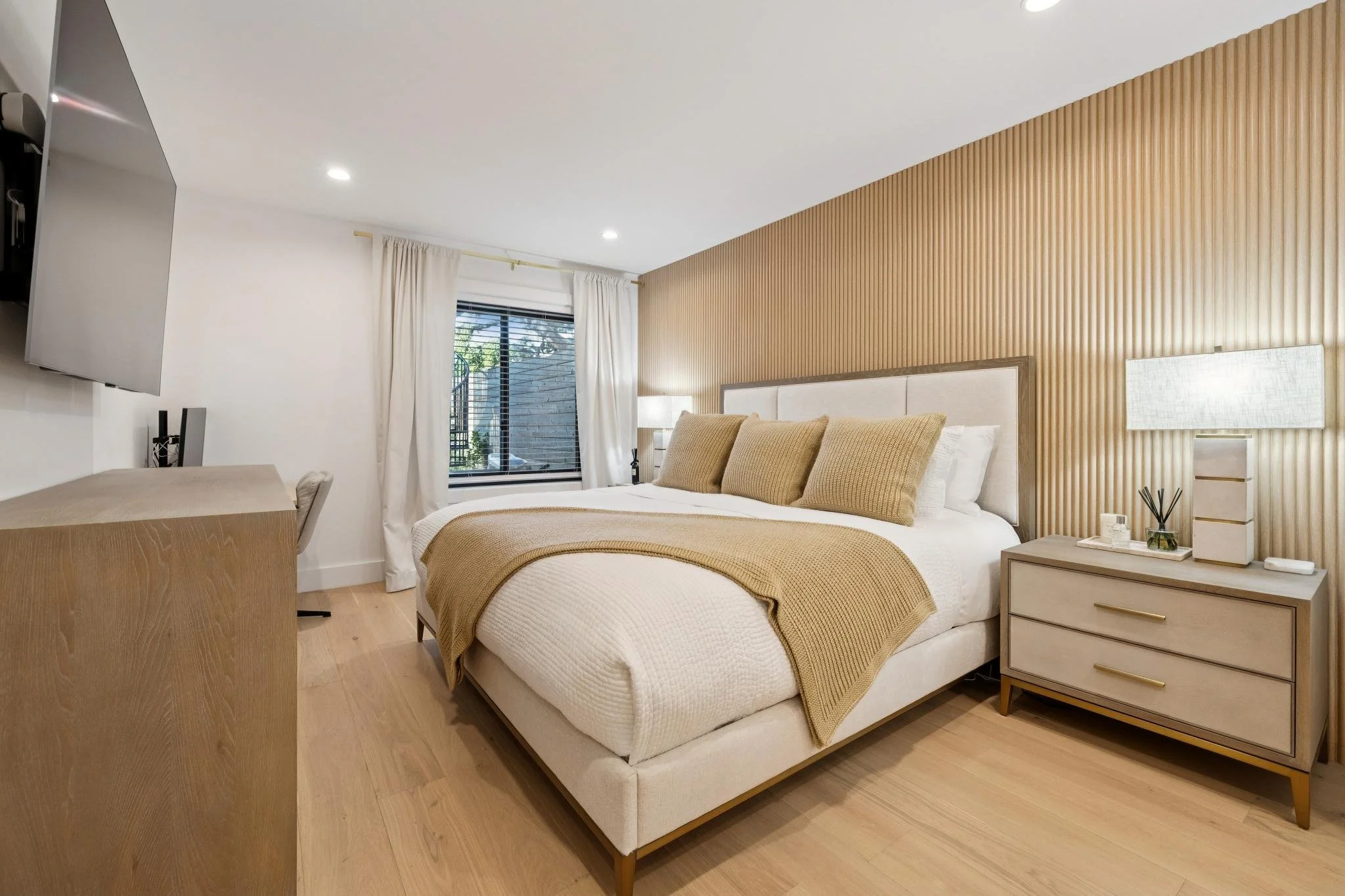 Modern bedroom with a bed, beige bedding, and pillows, wooden floor, nightstand with lamp, and window with white curtains.