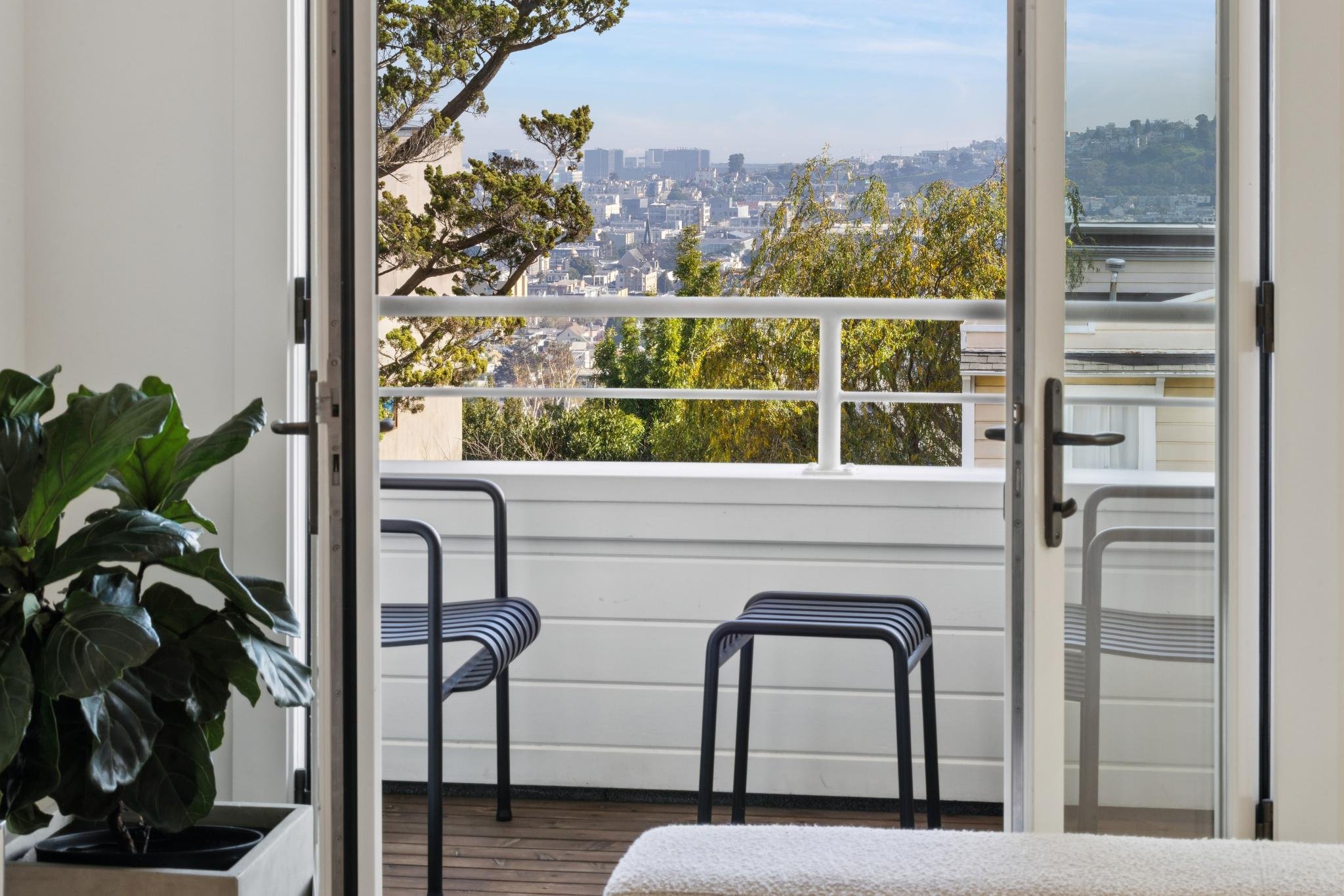 View from an open door leading to a balcony with two black chairs, a potted plant, and a cityscape with trees and buildings in the background.