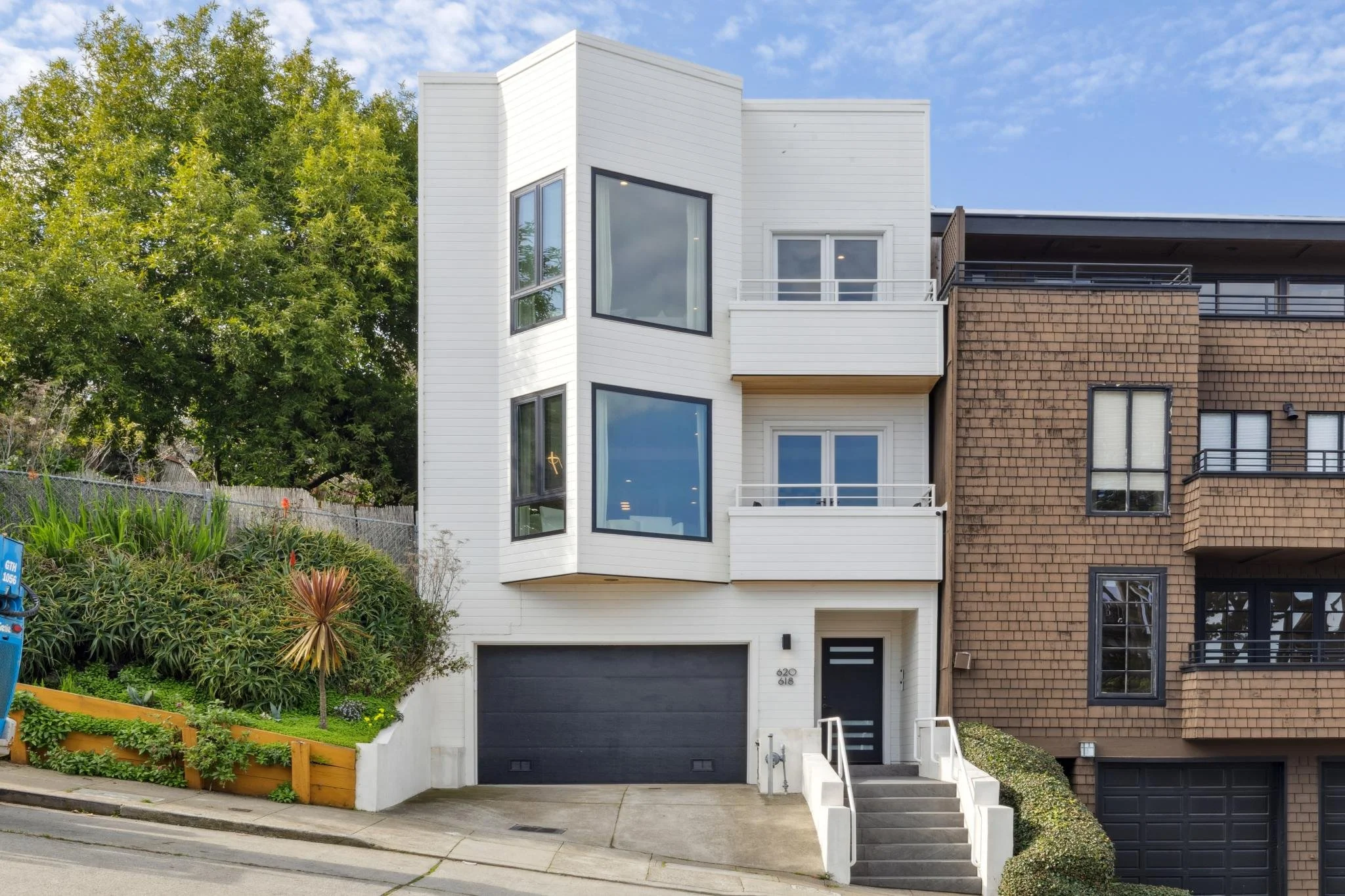 Modern white multi-story residential building with large windows, small balconies, and a garage, situated next to a building with a brown brick exterior. Green foliage and a sidewalk are in the foreground.