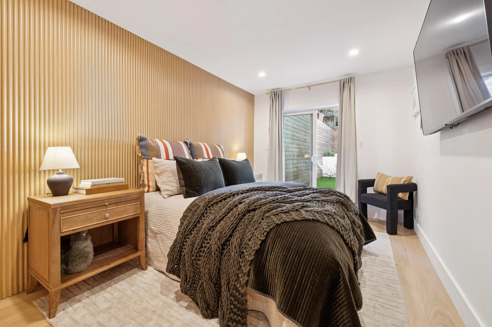 Bedroom with a bed, nightstand, lamp, and a sliding glass door leading to an outdoor space with a small patio and green lawn, decorated in neutral tones with a textured wood accent wall.