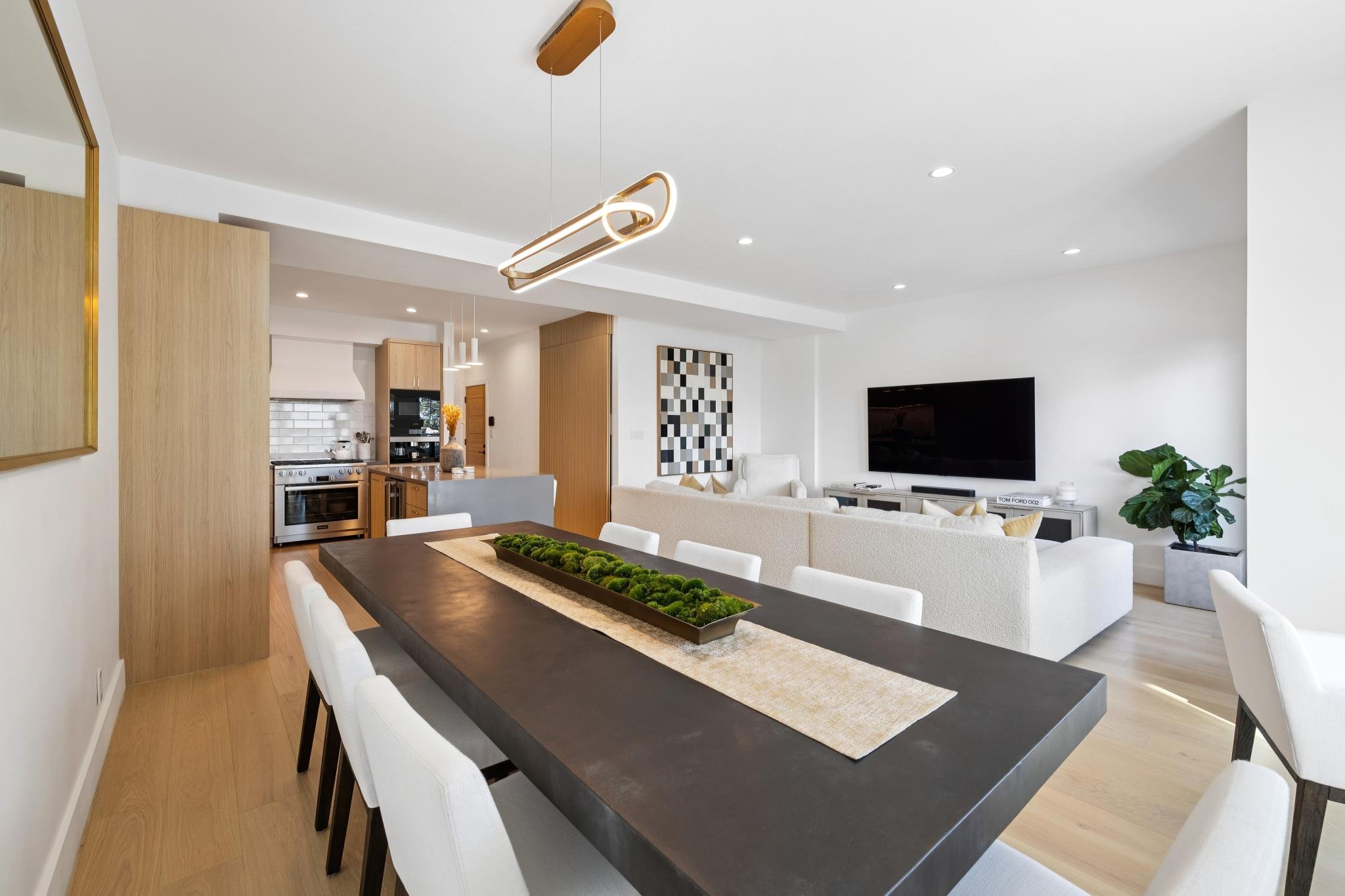 Modern open-concept living and dining area with a kitchen, white sofas, a large flat-screen TV, and minimalist decor.