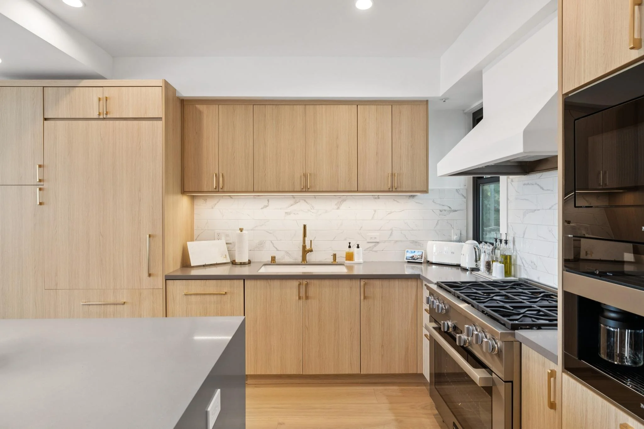 Modern kitchen with light wood cabinets, white marble backsplash, gray countertop, stainless steel stove, and black oven, with small appliances and a window.