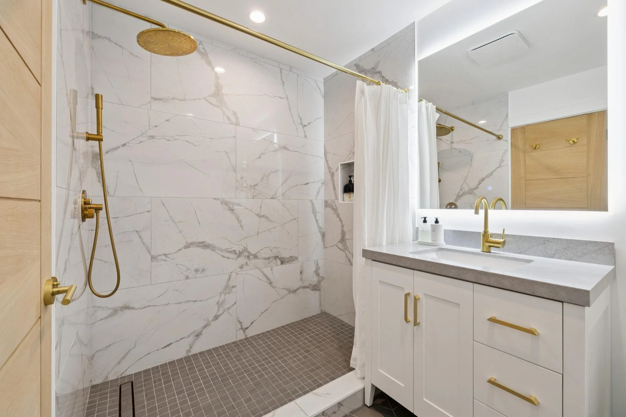 Modern bathroom with walk-in shower, white marble walls, gray tile floor, white vanity with gold hardware, large mirror, and gold fixtures.