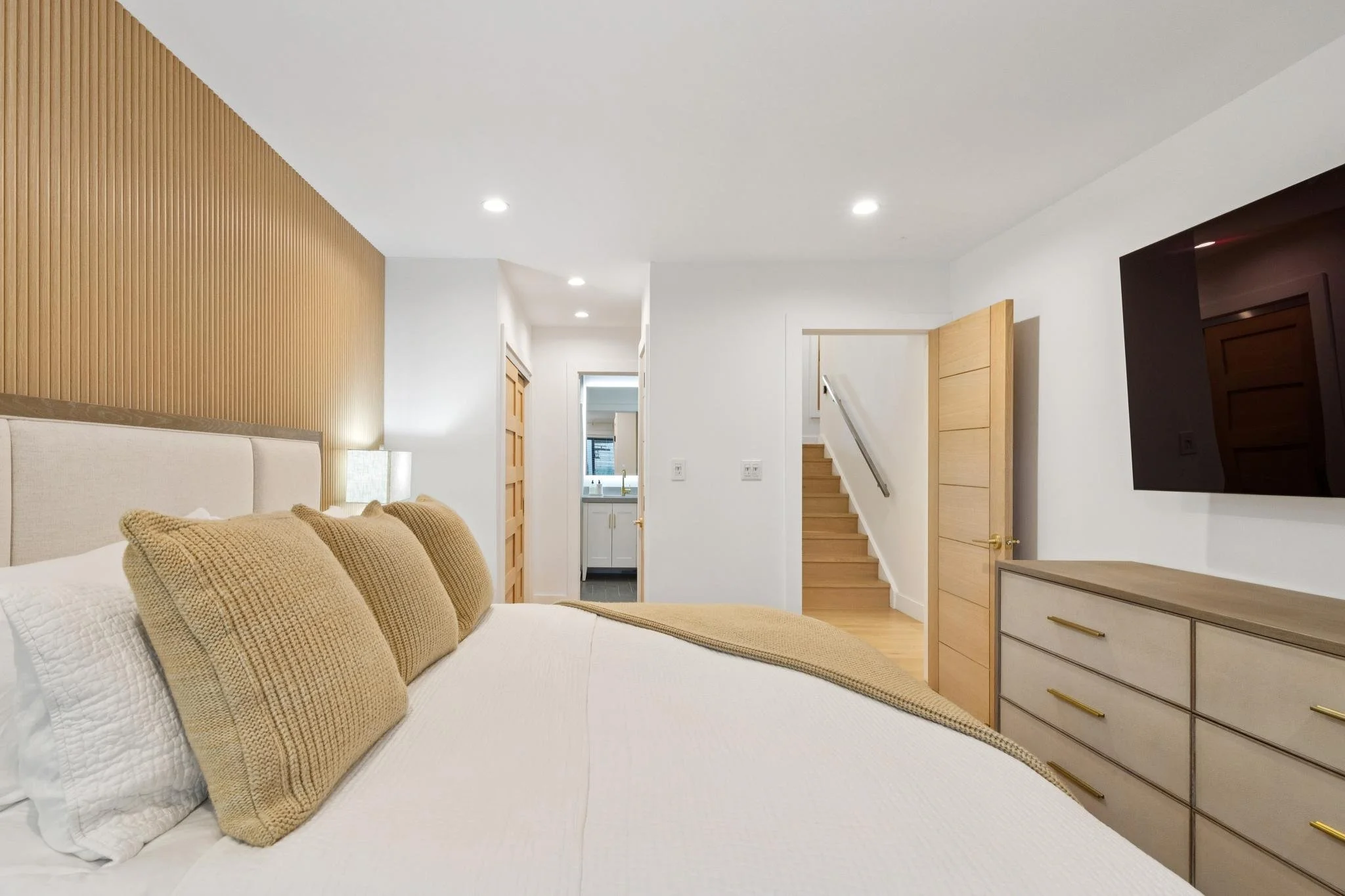 Modern bedroom with beige bed, wooden accent wall, dresser, TV, and staircase in the background.
