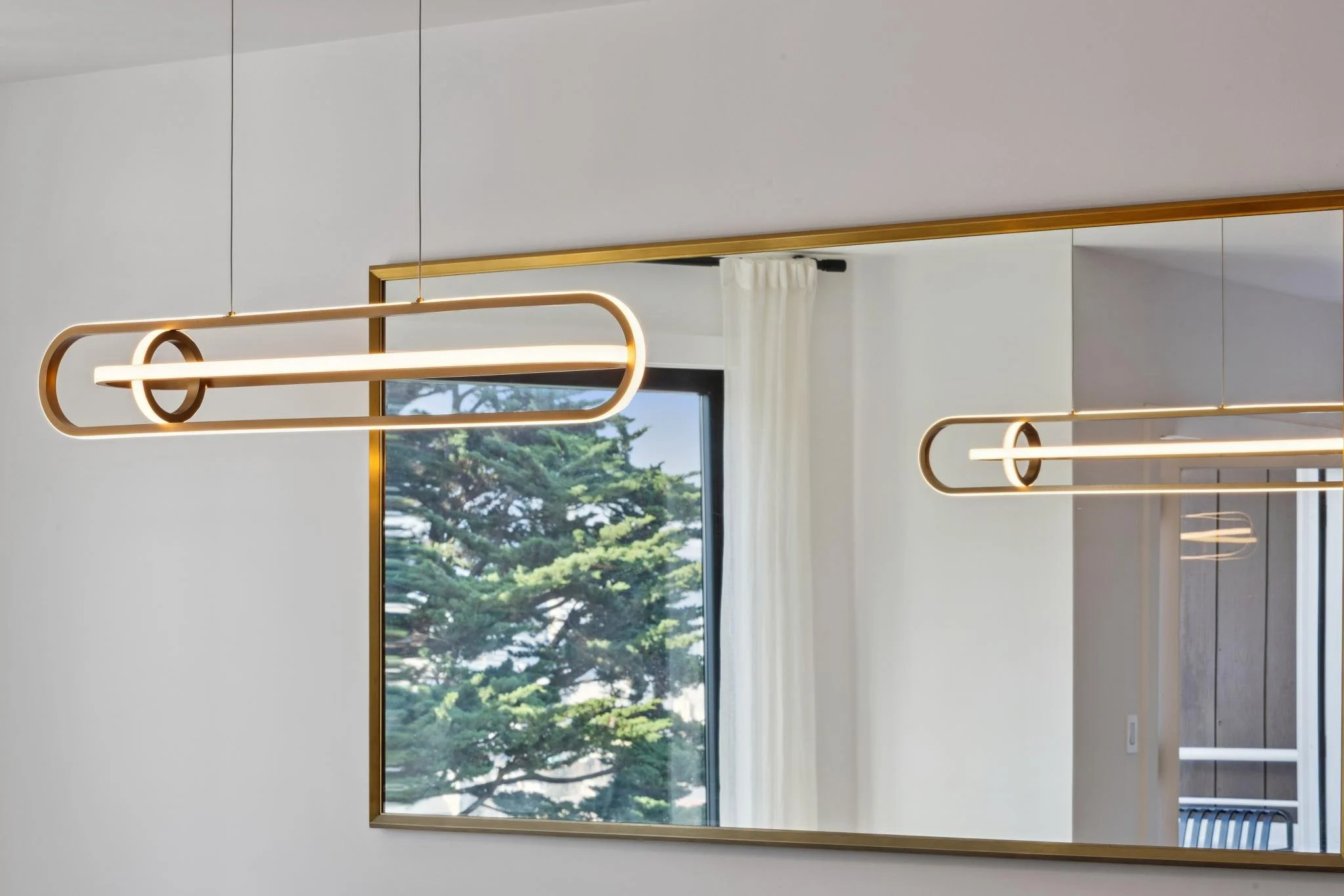 Modern minimalist pendant light fixtures with gold accents hanging in front of a large mirror, reflecting a window with a view of trees.