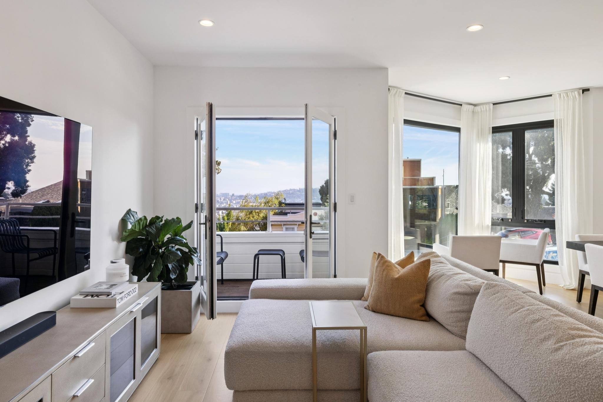 Living room with white sofa, beige pillows, glass doors opening to balcony, large windows with white curtains, and a view of city and trees outside