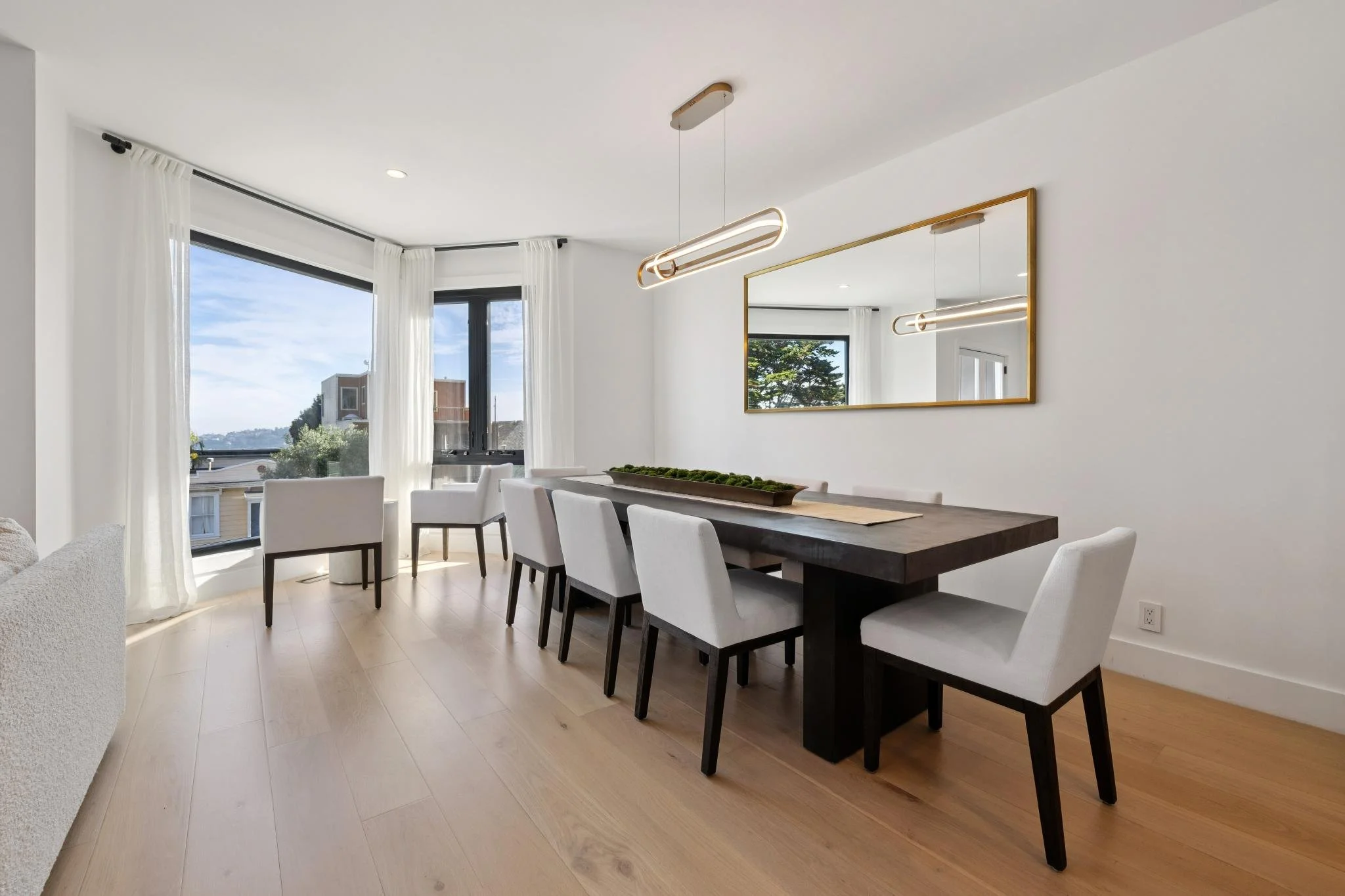 Modern dining room with a large dark wood table surrounded by white chairs, a large window with white curtains, a rectangular mirror on the wall, and a minimalist pendant light fixture.