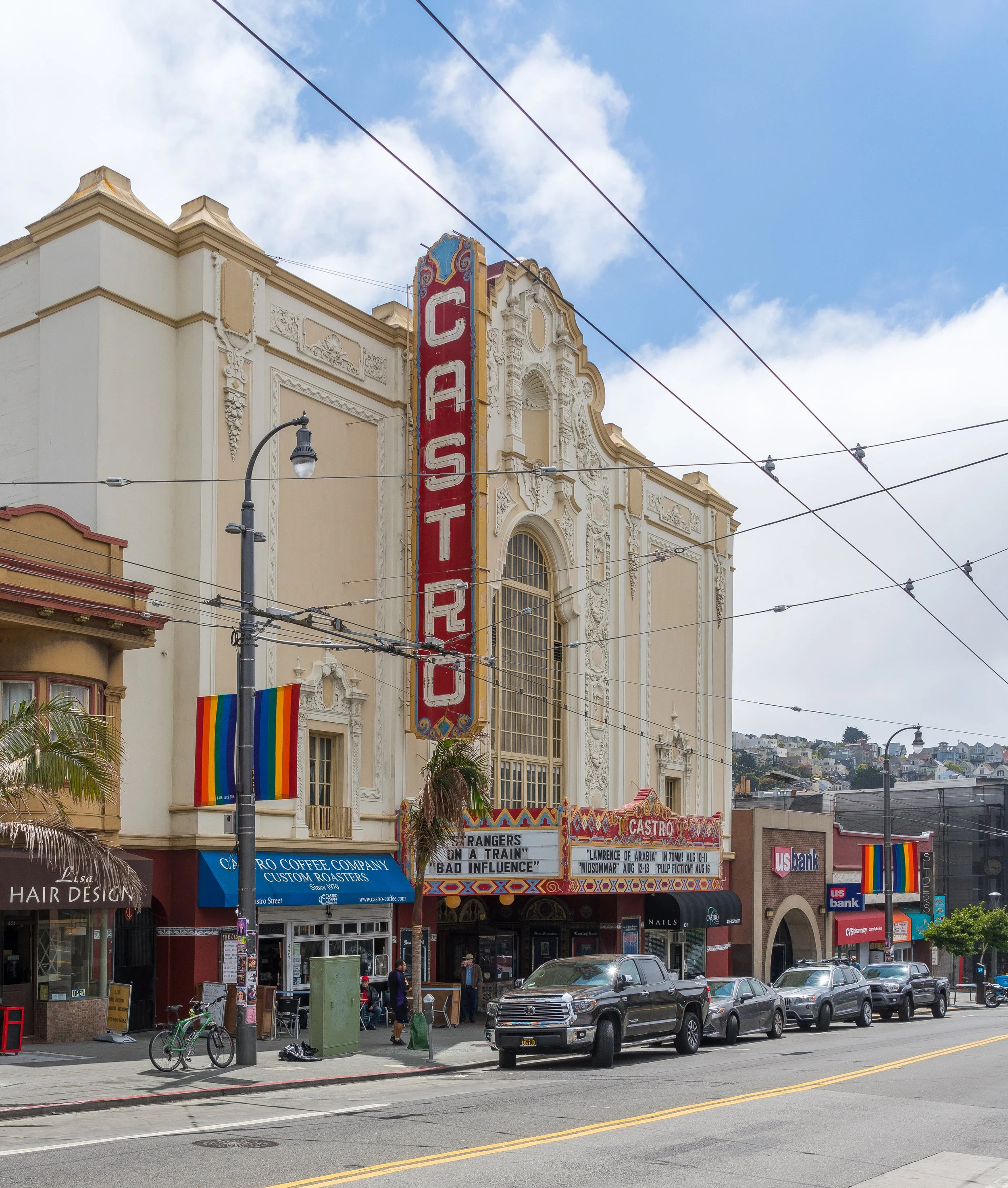 The Castro Theatre, a historic movie theater with a tall vertical sign reading 'CASTRO', is on a busy street with parked cars, pedestrians, and rainbow flags hanging. The marquee displays upcoming movie showings.
