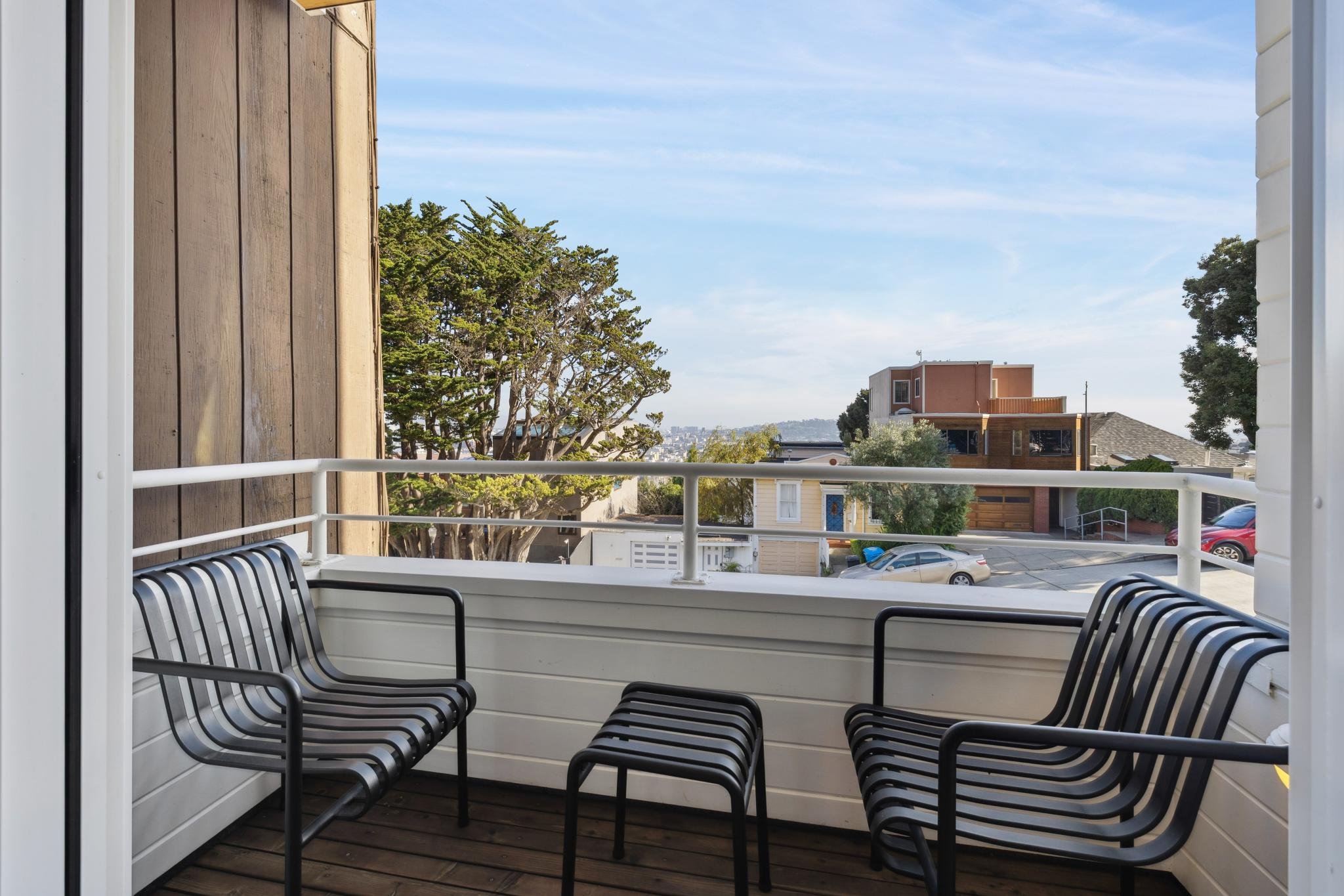 Balcony with two black metal chairs and stools, white railing, neighboring houses and trees, blue sky with clouds