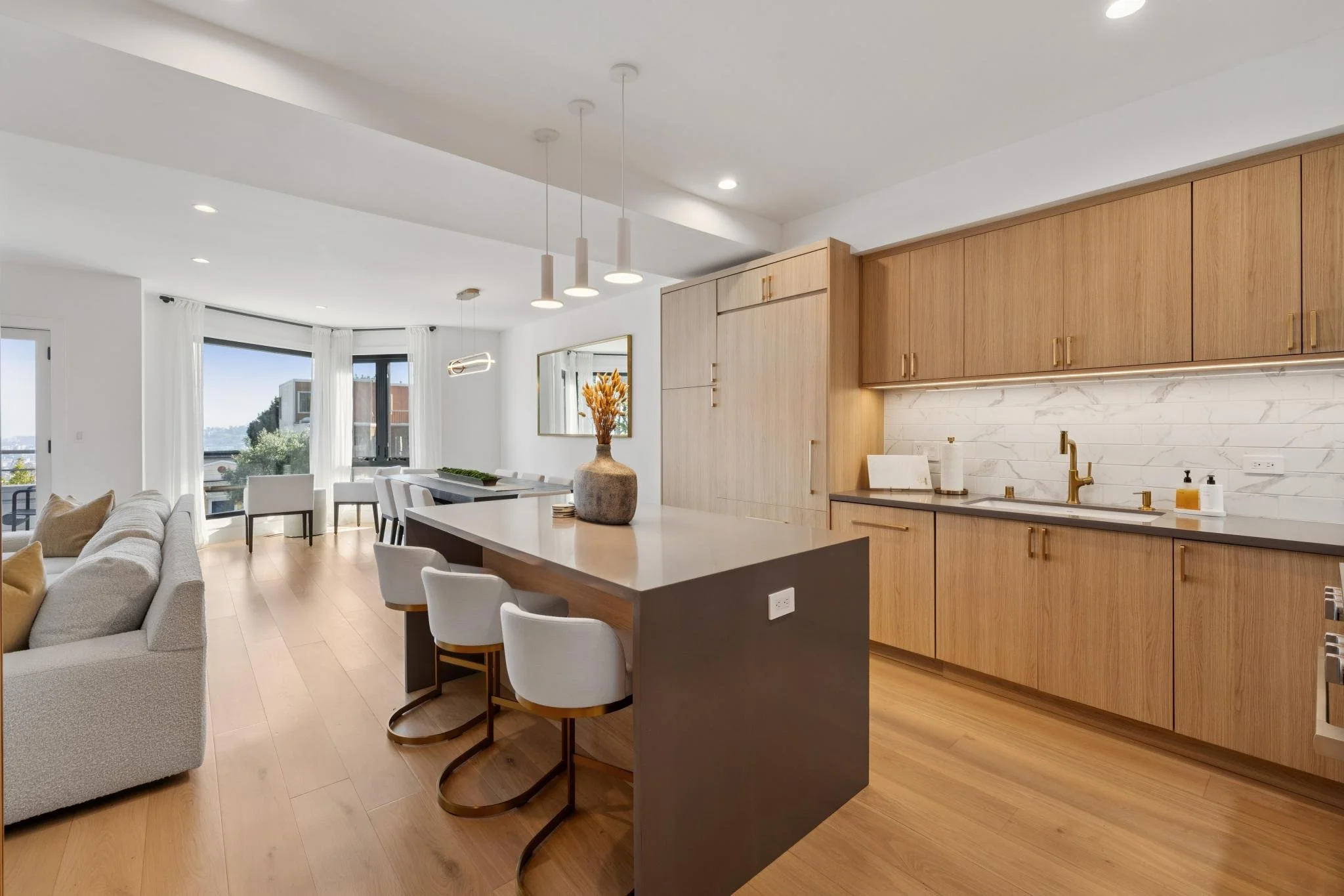 Open-concept living room and kitchen with wooden cabinets, white countertops, and a central island. Light-colored hardwood flooring, large windows with white curtains, and modern pendant lighting.