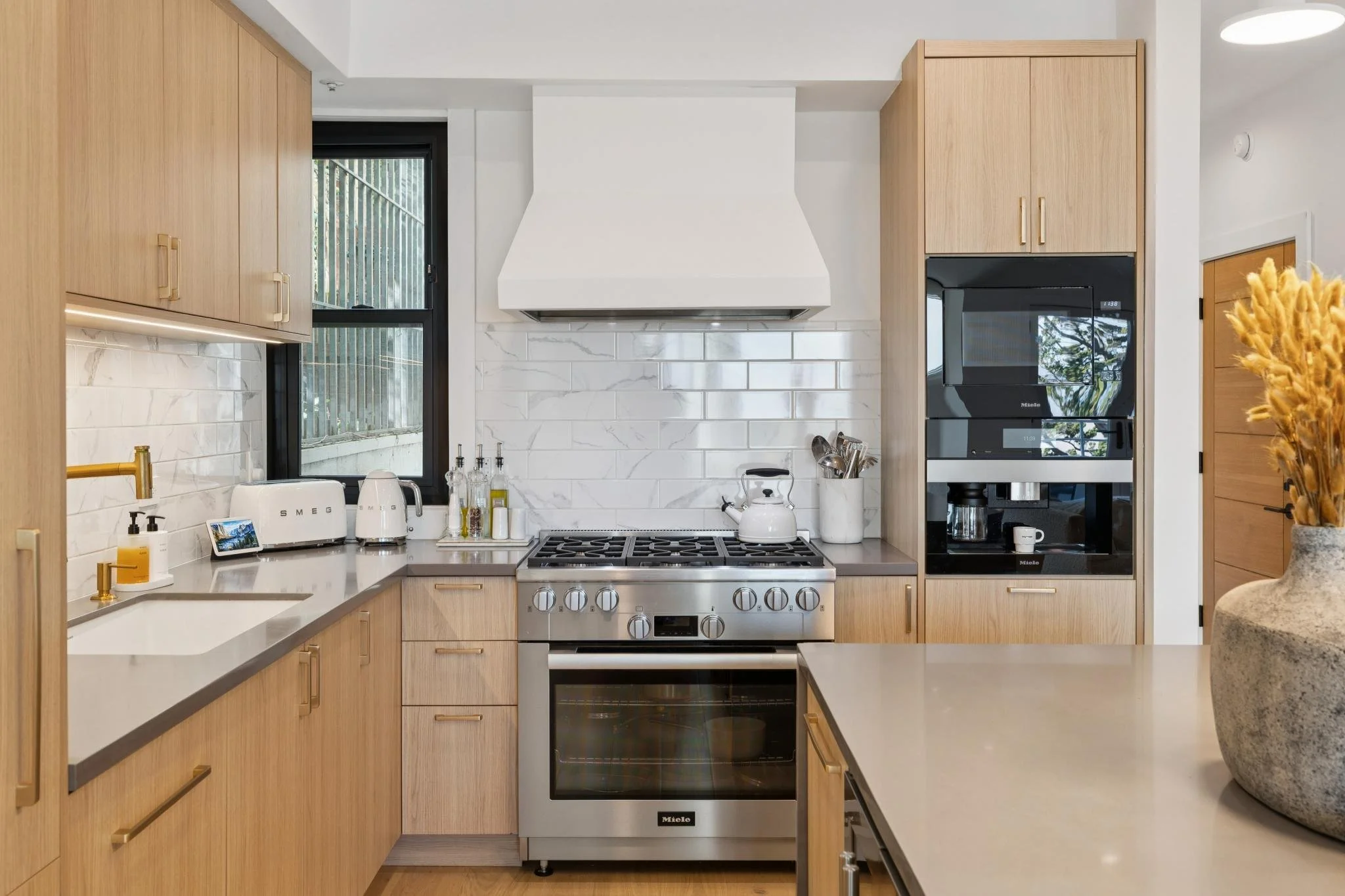 Modern kitchen with light wood cabinets, white backsplash, stainless steel stove, and appliances. A window with black frame is above the countertop.