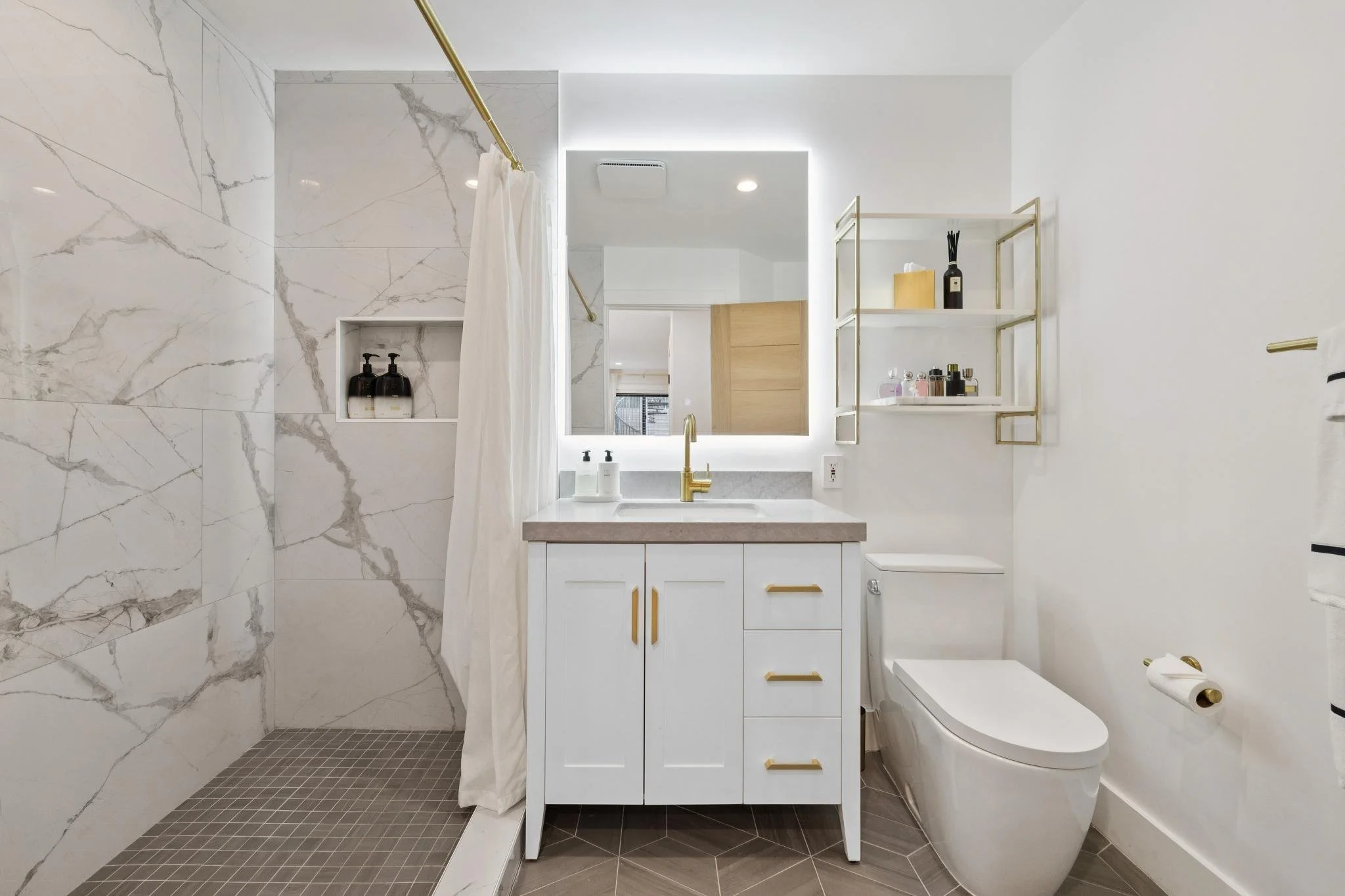 A modern bathroom with a white vanity, gold fixtures, a mirror with backlighting, a toilet, and a walk-in shower with marble tiles.